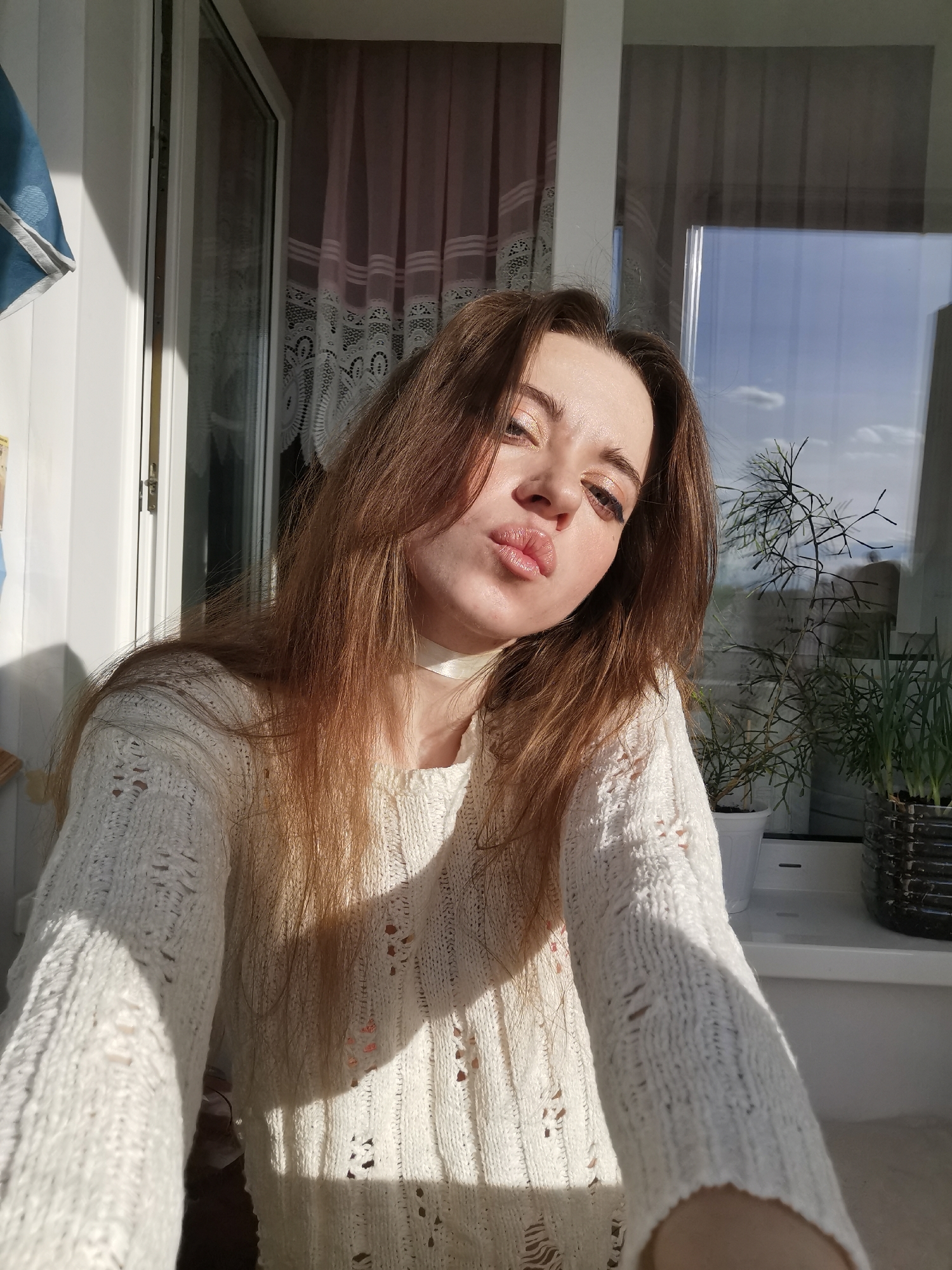 A woman with long brown hair and light skin is posing for a selfie indoors near a window. She is wearing a white, crocheted sweater and a white choker. Her lips are puckered as if giving a kiss. Sunlight illuminates her face and the sweater, creating shadows in the crochet pattern.
Behind her is a window with a view of a partly cloudy sky and some greenery. There are two potted plants on the windowsill. To the left, a slightly obscured window with light pink curtains is visible. A blue object hangs near the left window, partly out of frame. The location is Unknown, Unknown.