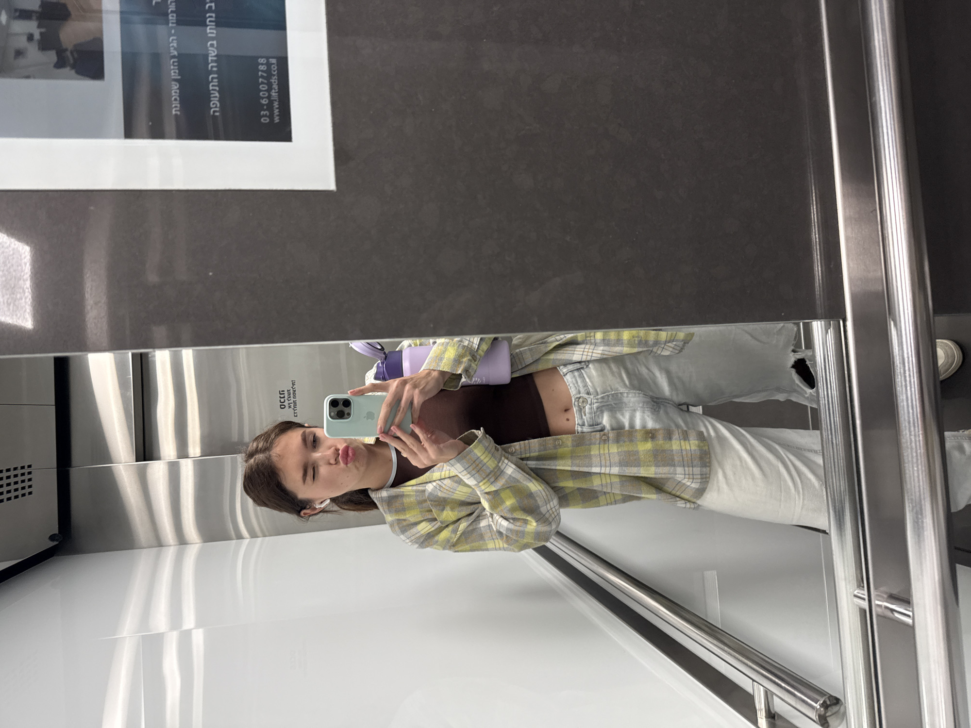 A woman takes a selfie in an elevator. The background features elevator walls, a handrail, and a reflective surface displaying a framed advertisement with text in Hebrew, including a phone number, 03-6007788. Based on the phone number and the Hebrew text, the location is likely Ramat Gan, Israel.