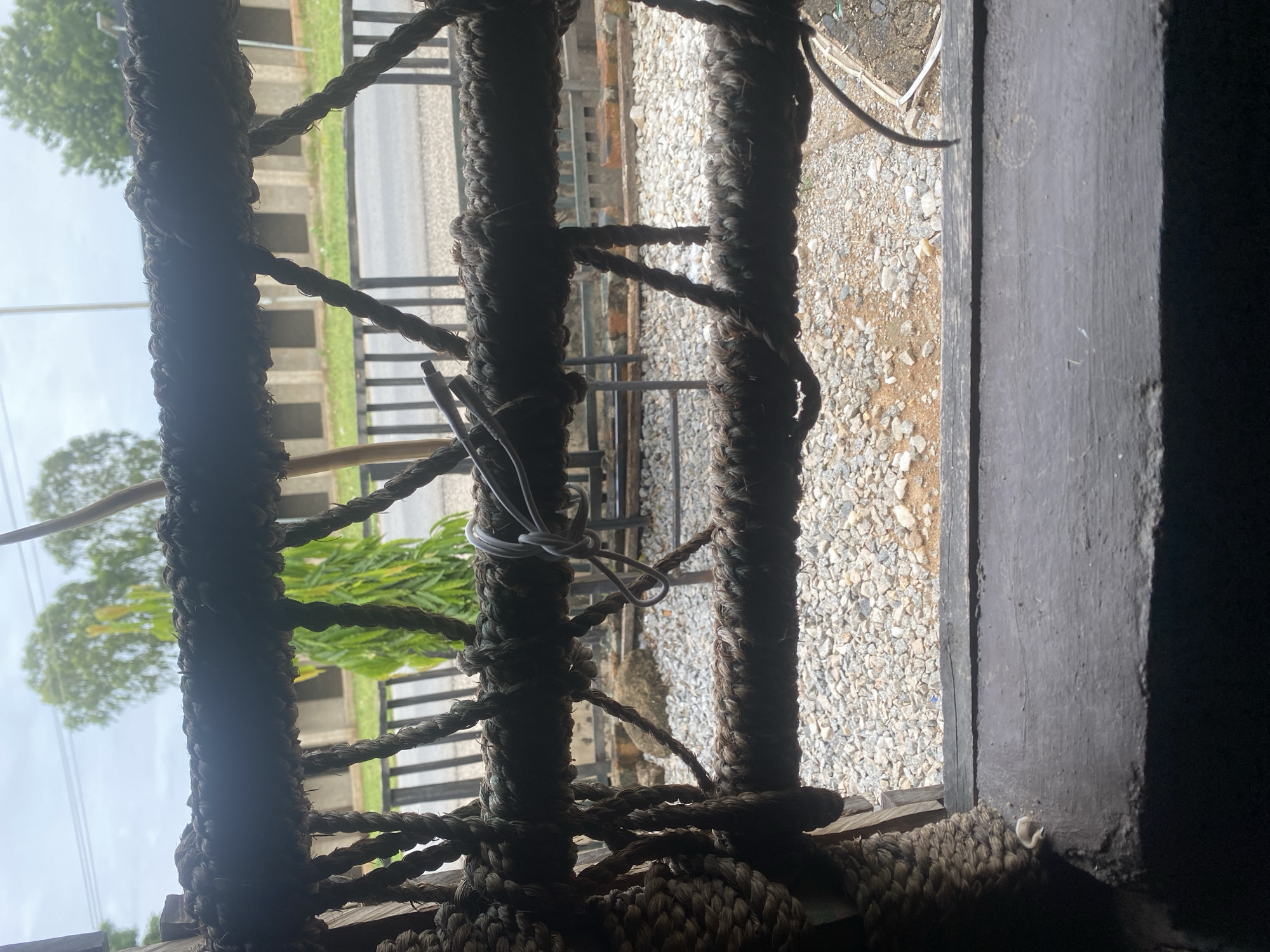 This is a close-up, rotated image of thick, dark ropes that appear to be part of a structure, possibly a hammock or a woven barrier. The ropes are twisted and sturdy, with some areas showing wear and tear. White rope is tied around one of the thick ropes.
Through the gaps in the ropes, a blurry background is visible. This background suggests an outdoor scene. In the distance, there are trees with green foliage and a cloudy sky. Below the trees, there is a glimpse of what looks like a road with some concrete structures bordering it. The ground area visible is covered with small, light-colored stones or gravel.
The image has a rustic, handmade feel due to the prominent ropes. The lighting suggests it might be daytime, possibly overcast or with diffused light, as there are no harsh shadows. The overall impression is one of being inside or looking out through a natural or crudely constructed barrier. Given the location context of Jalingo, Nigeria, the scene could be from a rural or semi-urban outdoor setting. No people or text are visible.