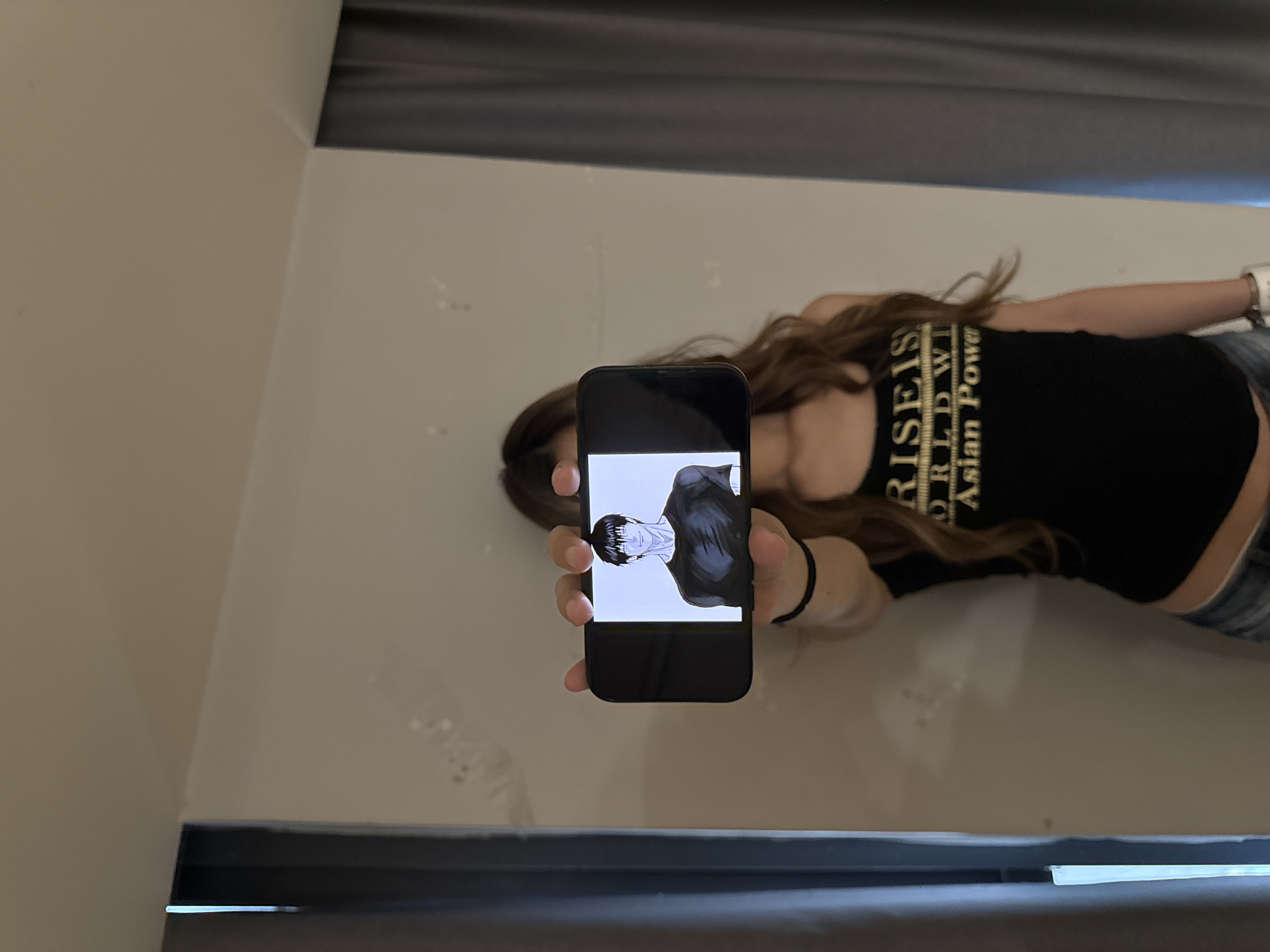 A woman is holding a black smartphone in her left hand, displaying an illustration of a man with black hair and a black shirt on the screen. The woman is wearing a black tank top with gold text that says "RISEIS WORLDWT Asian Power" and blue jeans. She also has a black bracelet on her right wrist and a white watch on her left wrist. She has long brown hair. The background consists of a white wall with a gray border at the top and bottom. The scene appears to be indoors. There is no indication in the provided image to confirm the location is in Liangjiang, China.