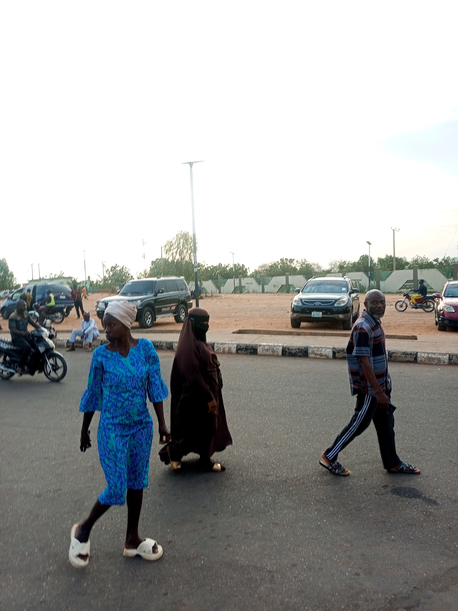 A street scene in Gombe, Nigeria, depicts several individuals and vehicles. A woman in a blue patterned dress and white sandals is walking in the foreground. Beside her is a woman in a brown niqab and gold sandals. A man in a striped shirt, black pants with white stripes, and brown sandals is walking to the right. Behind them are various cars and motorcycles parked or moving along the street. In the background, a low wall or barrier is visible. A streetlamp is also visible near the wall. Other pedestrians are visible on the sidewalks.