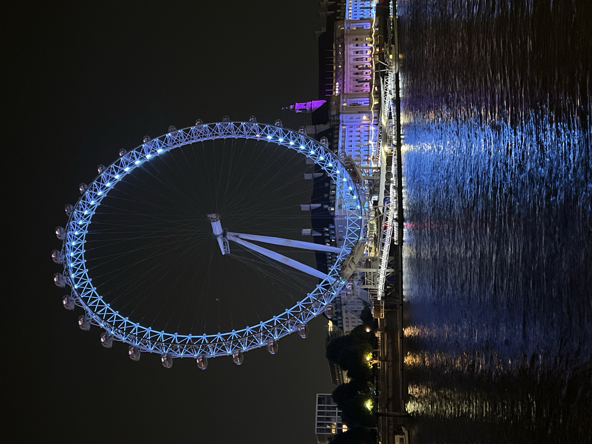 The image shows the London Eye Ferris wheel at night, illuminated in blue. The River Thames reflects the lights of the Ferris wheel and surrounding buildings, which include purple-lit structures. The sky is dark. Buildings line the opposite bank of the river.