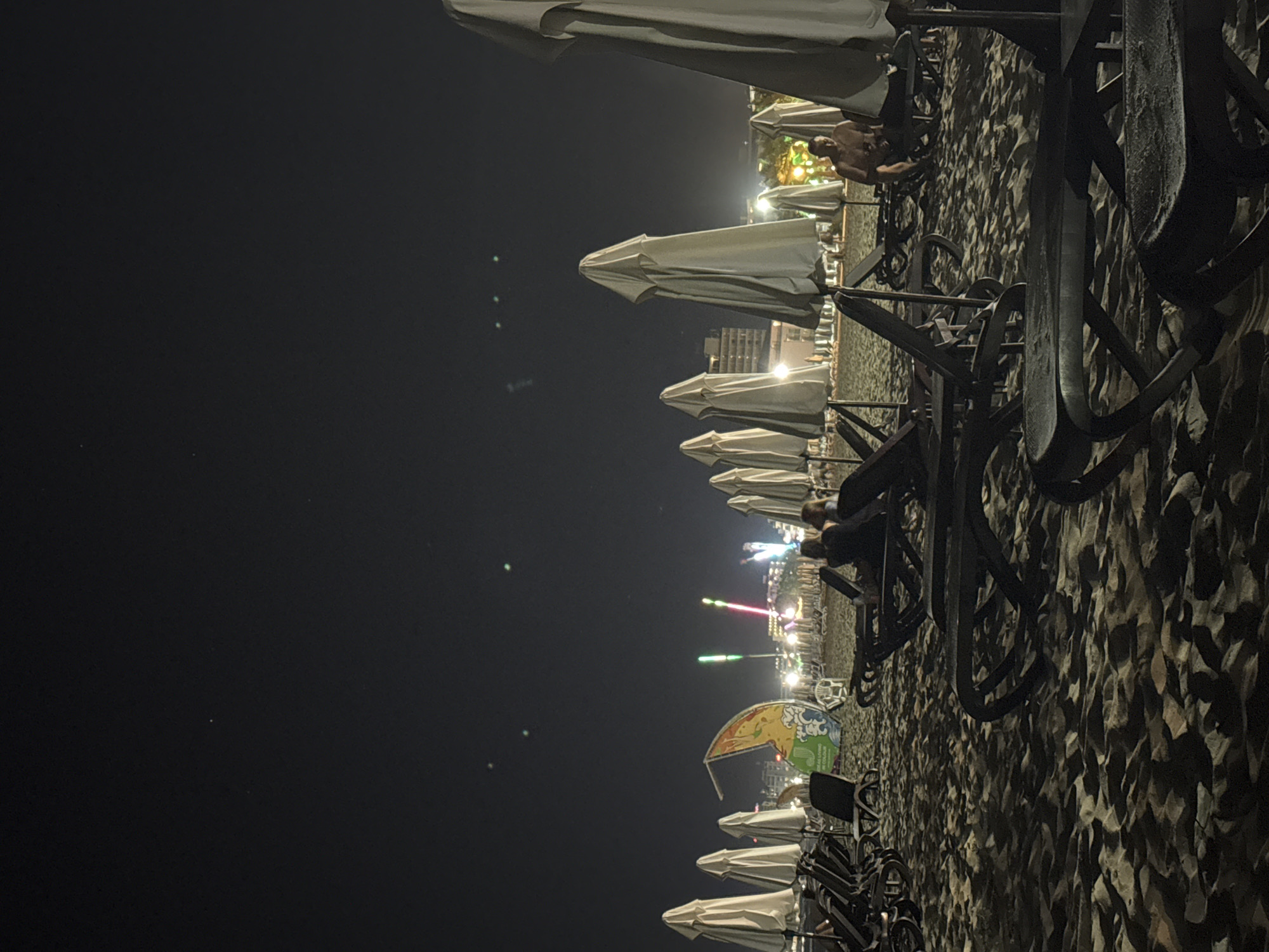 The image depicts a nighttime scene on a beach in Nesebar, Bulgaria. The foreground is dominated by rows of empty, dark-colored beach chairs and closed white umbrellas, arranged on sandy ground. In the mid-ground, several people are visible, some sitting in chairs and others standing. One man is seated on a lounge chair on the right side of the image, shirtless. Further back, the scene transitions to a more illuminated area with buildings and amusement park rides, indicated by brightly colored lights and structures. The sky above is dark with a few faint, greenish lights. The overall impression is of a quiet, post-dusk atmosphere on a beach that has transitioned from daytime activity to nighttime ambiance, with hints of a nearby entertainment district.