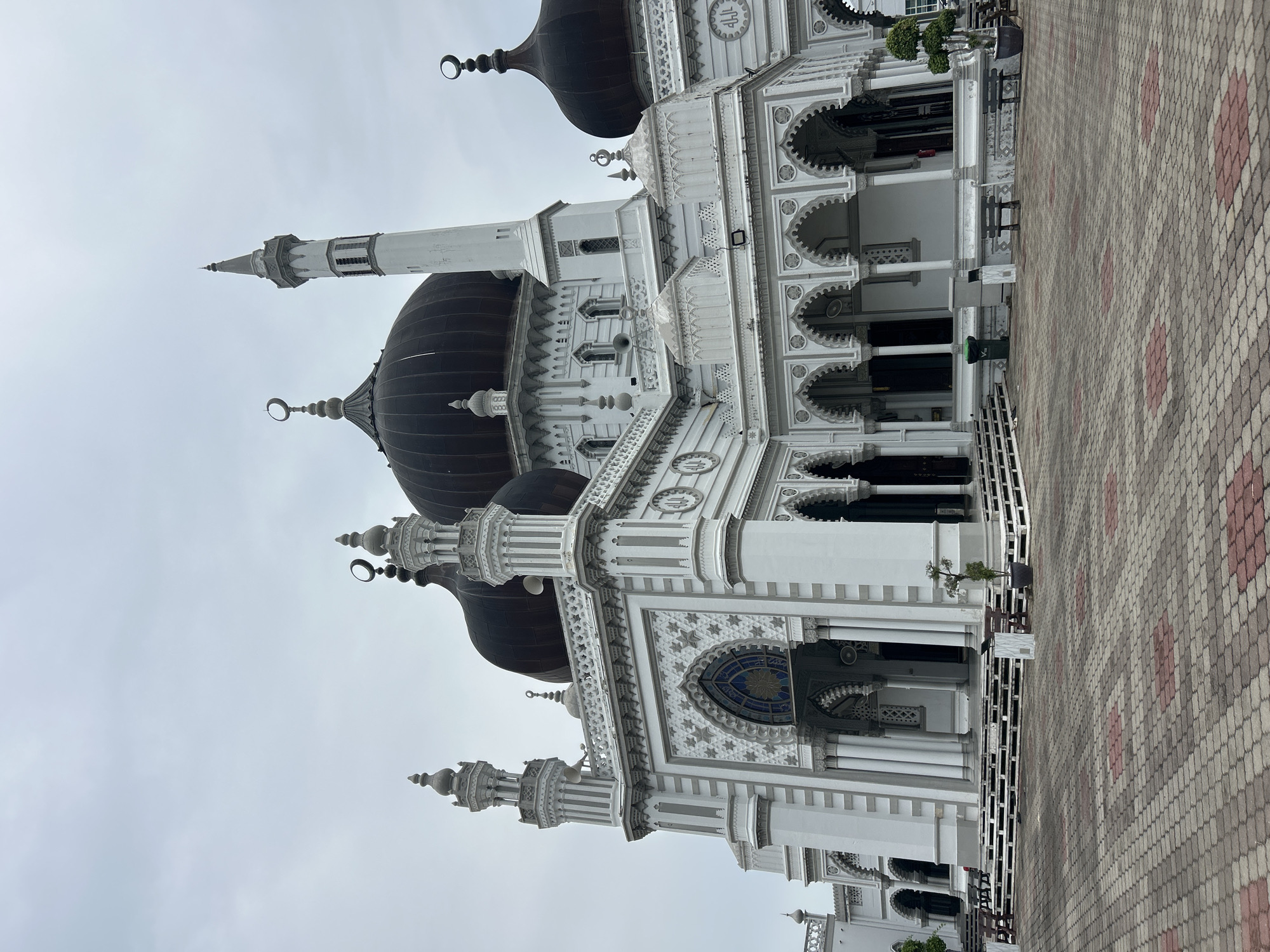 The image shows the Zahir Mosque in Alor Setar, Malaysia, under an overcast sky. The mosque is predominantly white with dark domes. It features intricate architectural details, including multiple minarets, arches, and decorative patterns. A paved courtyard with a geometric brick pattern is in the foreground. Benches and planters are visible in front of the mosque. No people are present in the image.