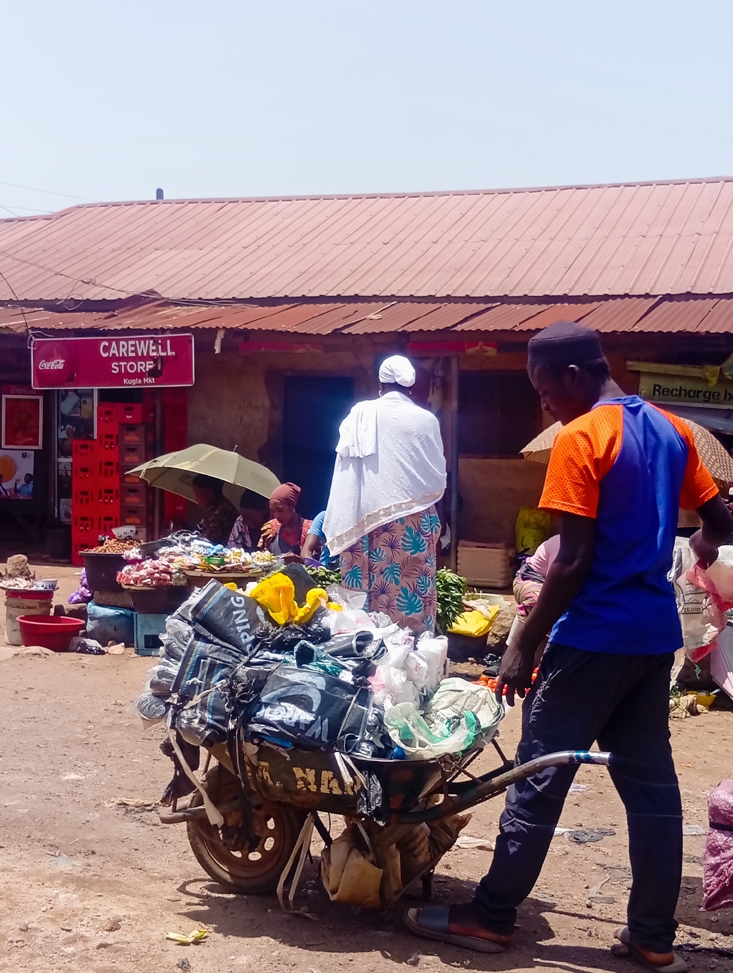 The image captures a bustling outdoor market scene in Bukuru, Nigeria, on a bright, clear day. The foreground is dominated by a man in an orange and blue t-shirt and dark pants, pushing a wheelbarrow loaded with an assortment of items, primarily wrapped in plastic bags and sacks. The wheelbarrow itself is old and worn, with visible rust and straps holding the cargo in place.
Behind the man, a woman in a white head covering and a patterned dress stands near a market stall. She is partially obscured by an olive-green umbrella, also under which other individuals are visible. The stall appears to be selling produce and packaged goods, with several large bowls filled with food items. To the left, a red Coca-Cola sign is prominently displayed on a building that also has a sign reading "CAREWELL STORE." Red crates are stacked near the store entrance.
The environment is dusty, characteristic of an unpaved market area. The background reveals more market structures with corrugated metal roofs, some appearing weathered. Sunlight casts shadows, indicating it's likely midday. The overall impression is one of active commerce and daily life in a Nigerian market.