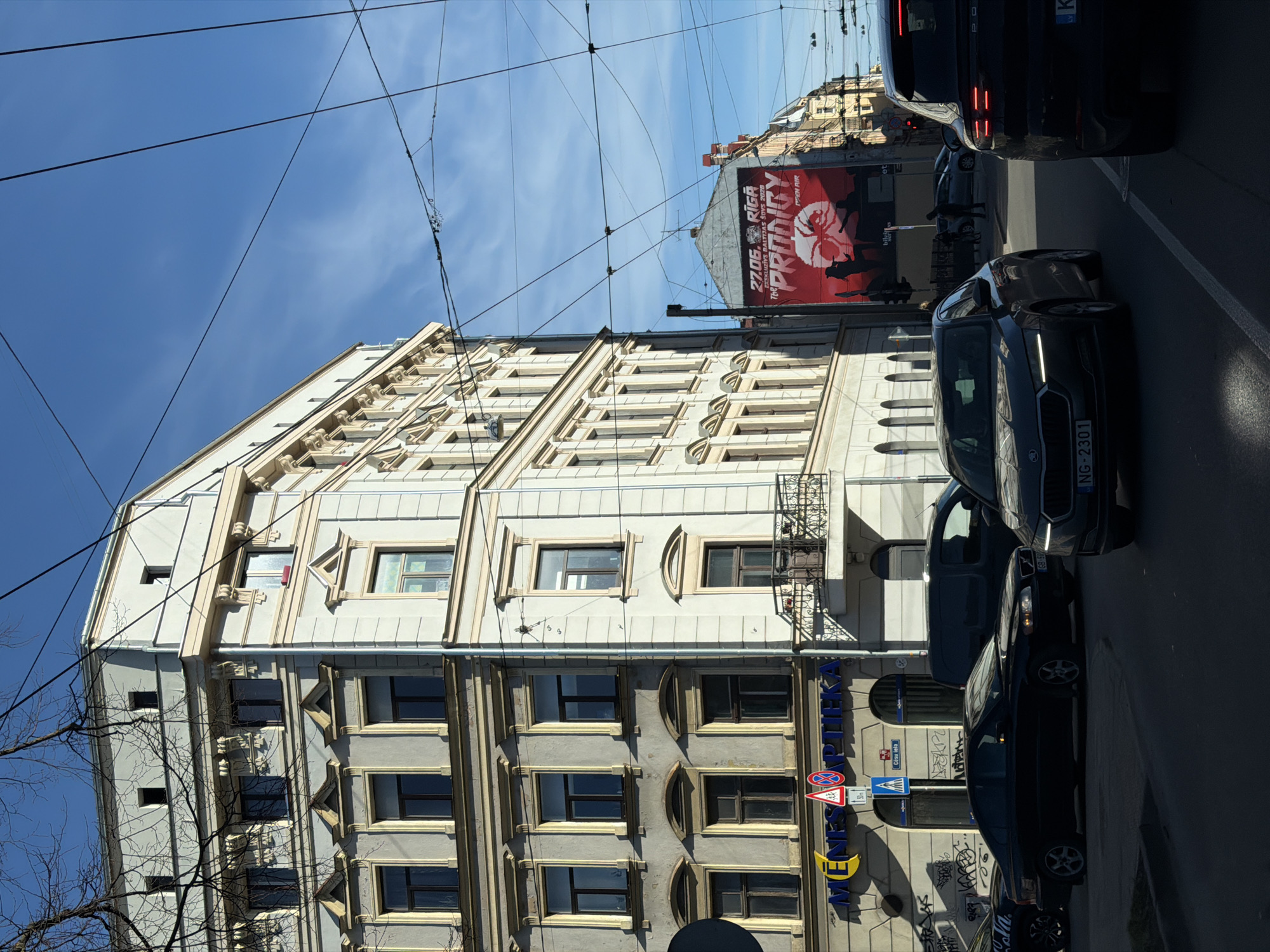 The image is a street-level perspective of a building in Riga, Latvia, on a sunny day. The building has a unique architectural style with contrasting textures and window designs on different floors. Power lines stretch across the sky above the building.
A large advertising billboard is affixed to another building in the background, partially obscured by other structures and power lines. The billboard advertises "The Prodigy" concert on 27.06 in Riga.
Several dark-colored cars are parked or driving along the street adjacent to the building. Street signs, including a crosswalk sign and a "no parking" sign, are visible near the building's base. Graffiti is present on the lower portion of the building.
The street address, "Cēsu iela 24B," is located on the side of the building, indicating its location in Riga. The car plates, specifically "GBS 487" and "NG-2301," can also be seen. The name "MENESS APTEKA" is visible as well.