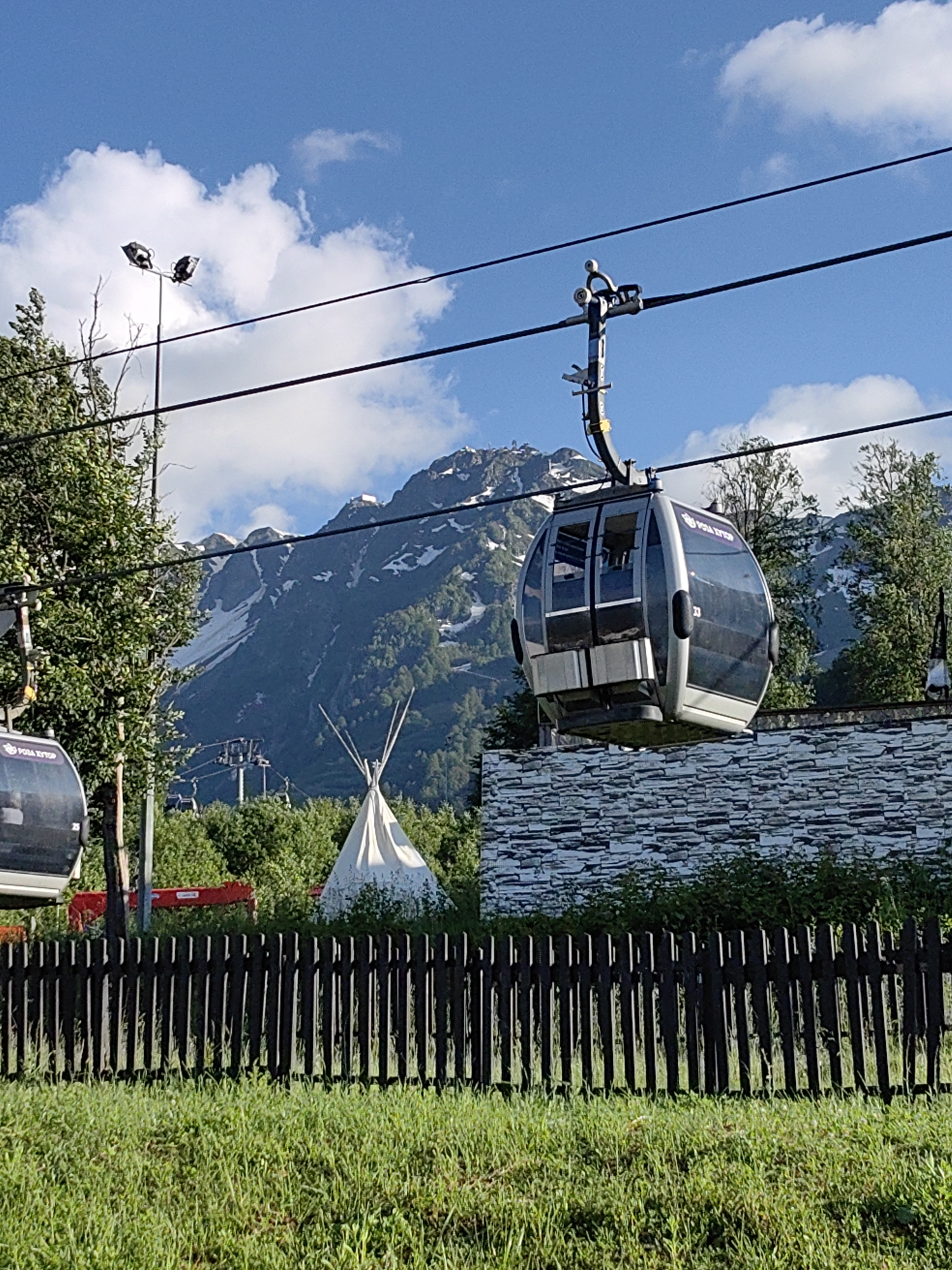 This media file captures a scenic view of a ski resort area on a sunny day, likely in the summer or early autumn. The main subjects are several gondolas of a ski lift traversing the frame, with one prominently in the foreground. The gondolas are modern, with tinted windows and an oval shape. The background features a majestic mountain range, partially covered in snow, suggesting a high-altitude location. Lush green trees and vegetation surround the base of the mountains and the resort area.
Notable details include a white teepee-like structure, hinting at a possible glamping or recreational area. A dark wooden fence runs across the lower part of the image, bordering a neatly mowed grassy area. The lighting indicates it is daytime, with bright sunlight casting shadows, suggesting a clear and pleasant weather. The sky is a vibrant blue with scattered white clouds.
There is some visible text on the gondola in the foreground: "РОЗА ХУТОР" (Rosa Khutor), which is a well-known ski resort in Estosadok, Russia. Another smaller number "33" is also visible on the gondola. The overall mood is peaceful and suggests an inviting outdoor recreational setting. No people are clearly visible within the gondolas or the immediate scene, but the presence of the ski lift implies people are either using it or have used it recently.