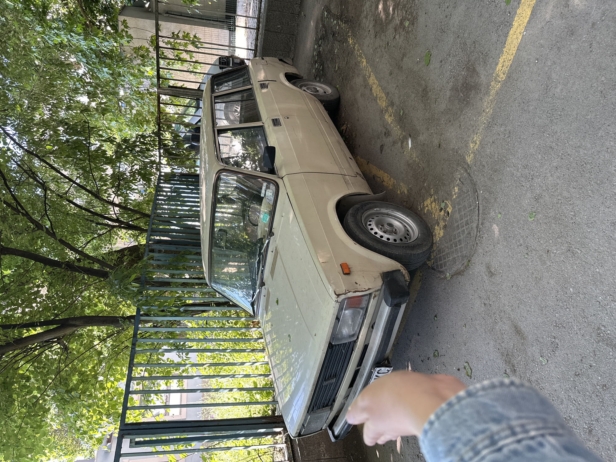 A vintage, beige Lada estate car is parked in an outdoor lot in Varna, Bulgaria. The car is dusty and shows signs of age, with some rust visible on the body. A metal fence, painted teal, borders the parking area, with lush green trees and foliage visible beyond it. Sunlight filters through the leaves, casting dappled shadows on the car and the asphalt ground, which is marked with faded yellow parking lines. A partially visible hand, wearing a denim jacket, is in the foreground, suggesting someone is taking the photograph. The car is facing to the right, and its rear is towards the fence. A manhole cover is visible on the ground near the front right wheel of the car. The overall impression is one of a quiet, urban setting, perhaps a residential area or a less-trafficked parking space.