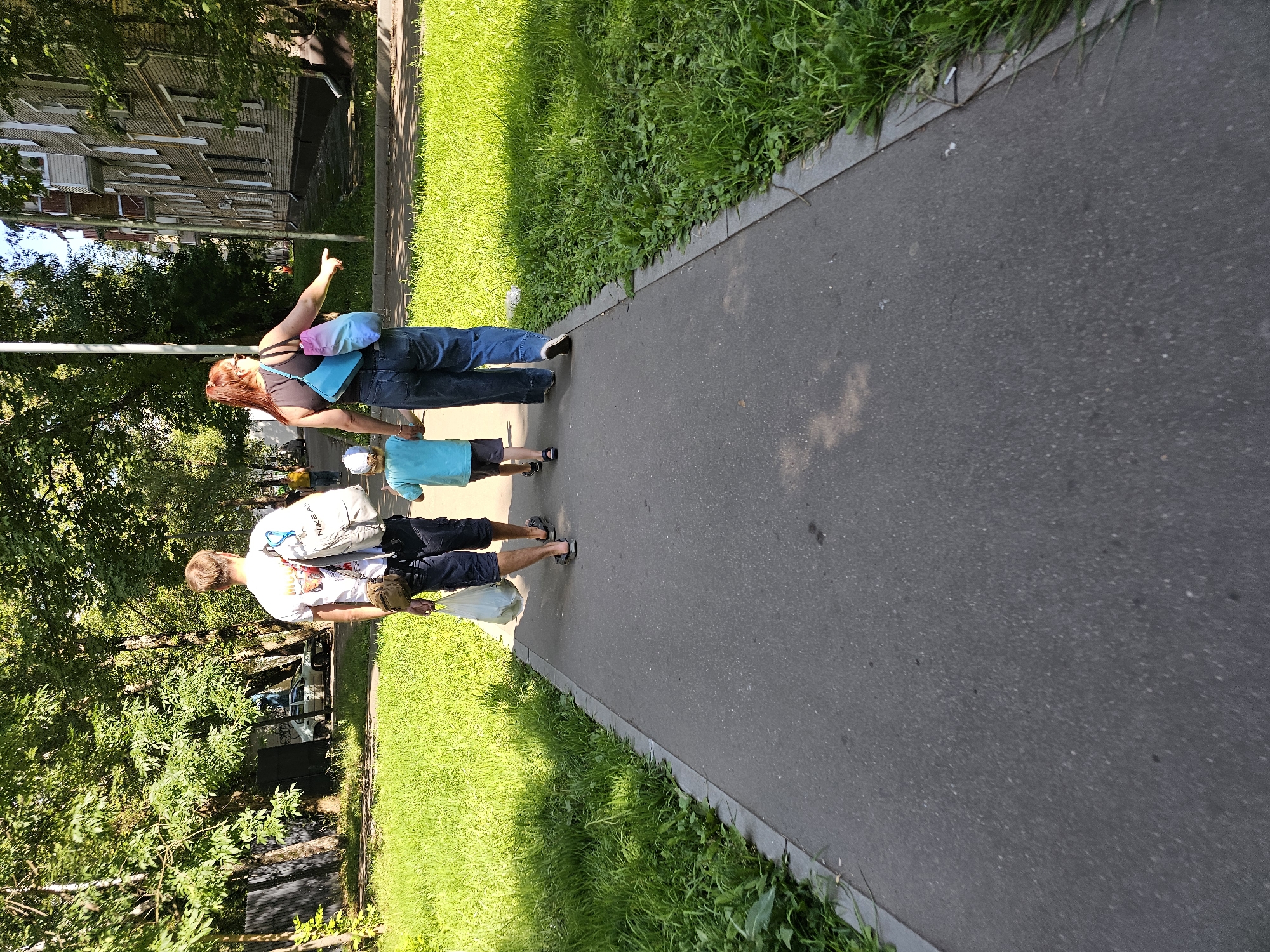 Three people, two adults and a child, walk away from the camera on a paved sidewalk. The adults are identified as a man and a woman. The man is wearing a white t-shirt, dark shorts, and sandals. He carries a white plastic bag in his right hand and a backpack on his shoulders. The woman is wearing a dark tank top, blue jeans, and sandals. She has a multicolored backpack on her shoulders and is pointing with her right hand. The child, in a blue shirt and shorts, walks between them. The sidewalk is bordered by a strip of green grass and trees. In the background, a multi-story brick building is visible on the right, and trees and a fence are on the left. The scene appears to be a residential area in Khimki, Russia, on a sunny day.