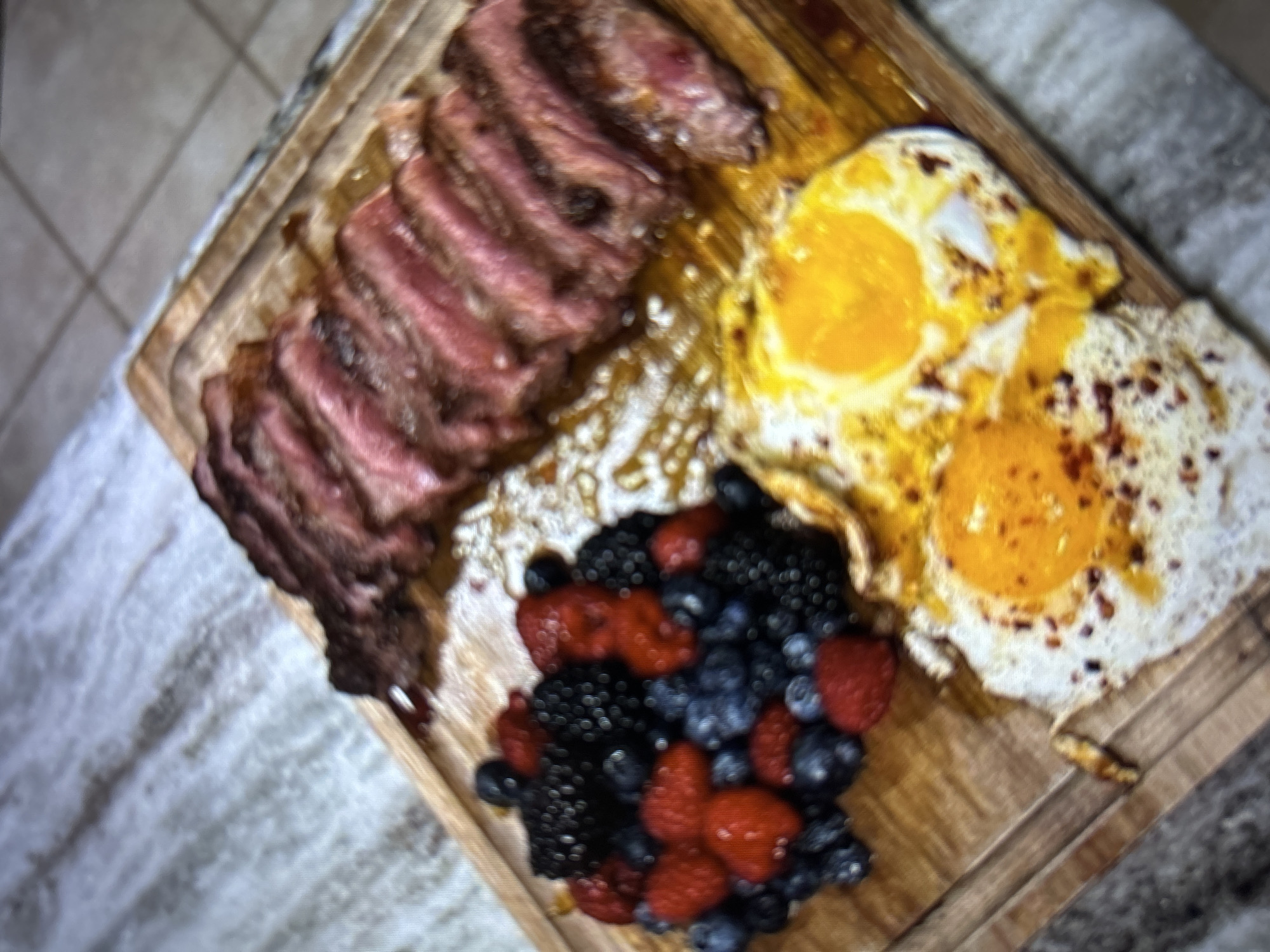 A close-up, overhead shot captures a wooden cutting board laden with a meal. Sliced steak, cooked medium-rare with visible pink interiors and seared edges, is arranged in a row on the upper right. To the left, a generous portion of mixed berries, including blackberries, raspberries, and blueberries, is piled high. At the bottom of the board, two fried eggs, their yolks still bright yellow and runny, are seasoned with what appears to be red pepper flakes. The food is presented on a light-colored, marbled surface that could be a countertop. There are no people or specific locations in Burgas, Bulgaria evident in this image.