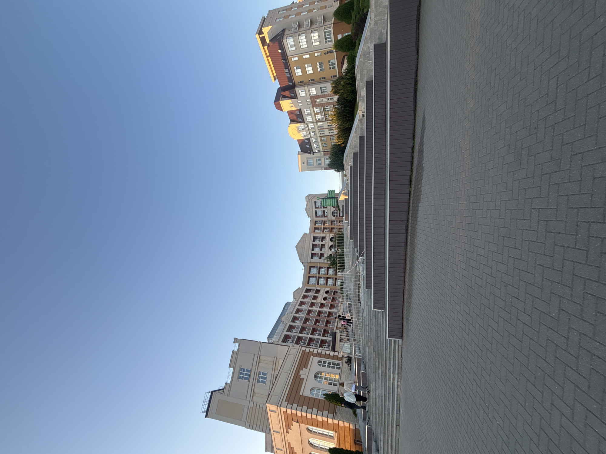The image depicts a street scene in Varaksino, Russia. Buildings are visible on both sides of the frame, with a paved walkway in the foreground. A set of steps runs parallel to one side of the buildings. Several people are seen on or near the steps, including two individuals who are walking. The sky is clear and blue. The buildings appear to be residential or mixed-use structures.