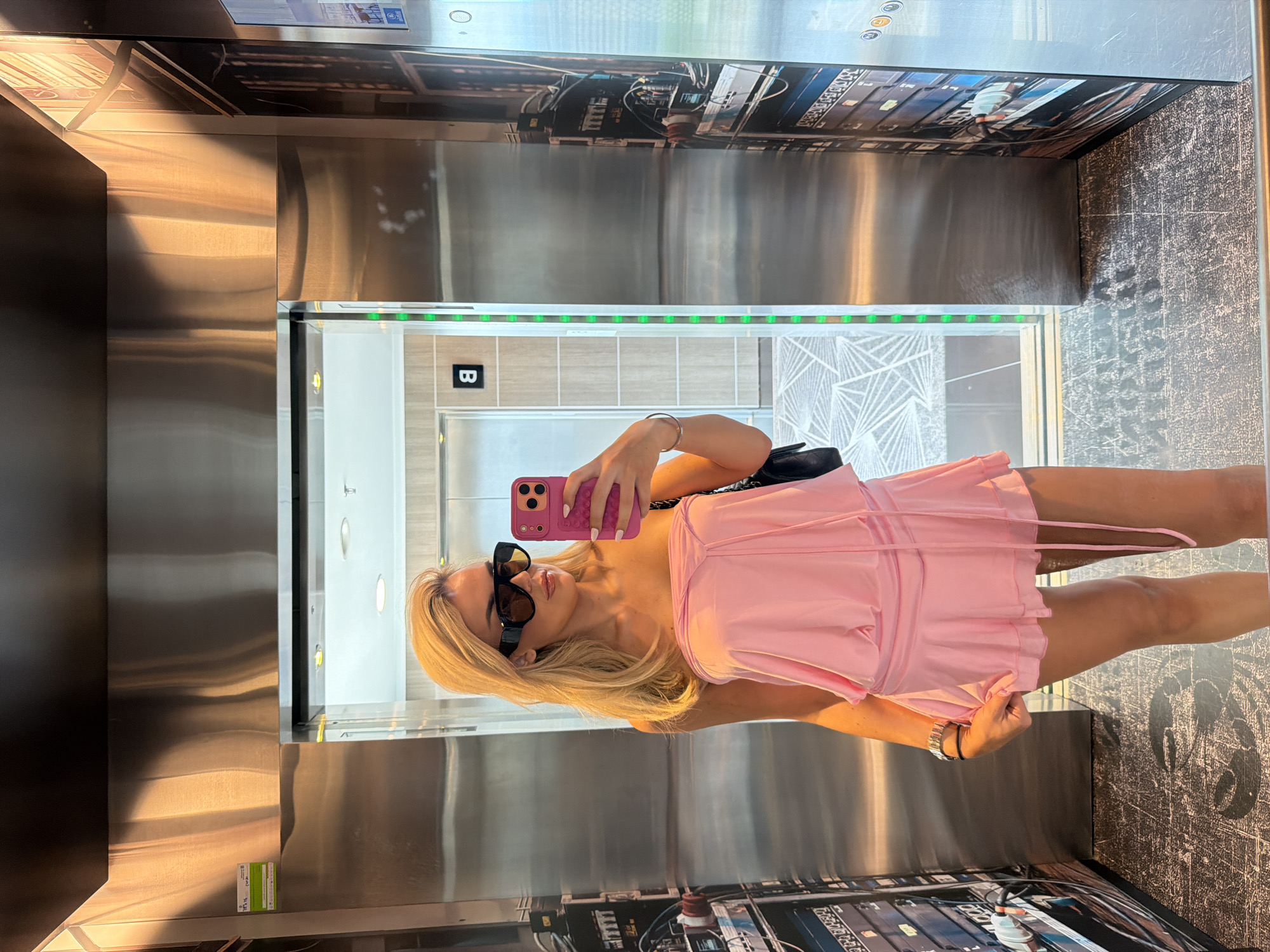 A blonde woman, wearing large dark sunglasses and a light pink strapless top, takes a selfie reflection in an elevator mirror. She holds a pink smartphone with a textured case in her right hand, positioned towards the mirror. A silver bracelet is visible on her left wrist, and a black bag strap crosses her right shoulder. The elevator interior features brushed stainless steel panels, with the right wall adorned by industrial-themed graphics depicting complex wiring and equipment, on which a "Hilton" logo is discernible. The floor of the elevator is dark with a textured, abstract pattern. Reflected in the mirror are exterior elevator features including a call panel with a black "B" button and a vertical strip of green indicator lights. Beyond the elevator, a modern hallway with light-toned walls and a distinct geometric floor pattern is visible. The scene is located within an elevator of a Hilton property in the city of Barcelona, Spain.
