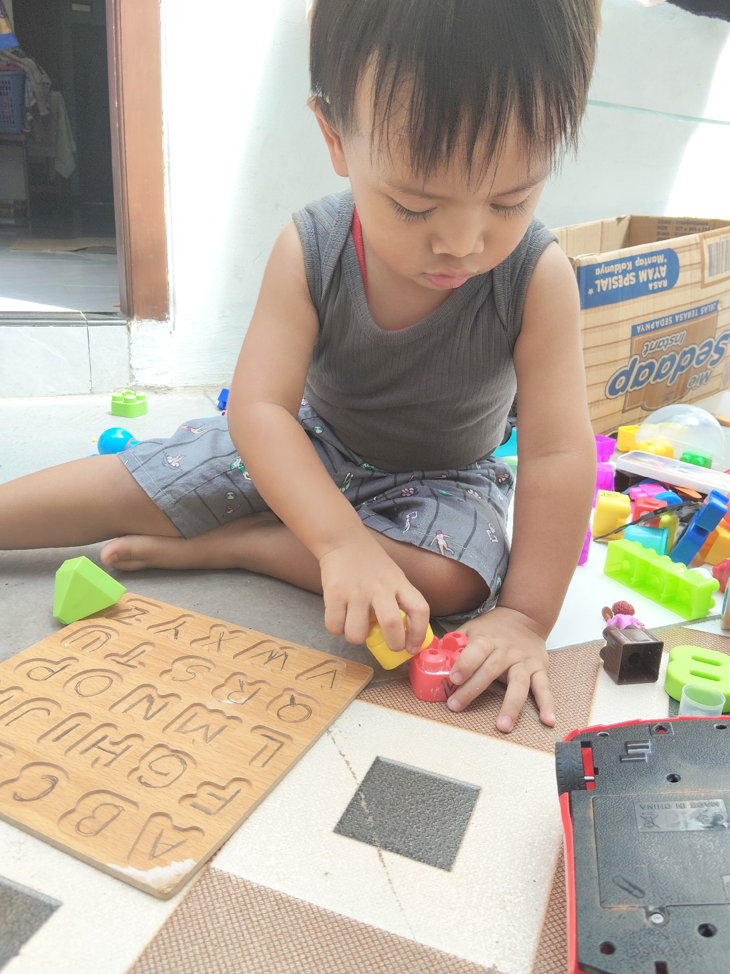 A young child is sitting on the ground, playing with toys. The child is wearing a gray tank top and gray shorts with a pattern of white figures. In front of the child is a wooden alphabet puzzle with the letters A through Z carved into it. The child's hands are engaged with a red and yellow toy. Scattered around the child are various colorful building blocks and other small toys, including a small brown figurine of a person and a red toy car. In the background, a cardboard box with Indonesian text, possibly food packaging, is visible. The scene appears to be outdoors, near a doorway with a tiled floor. The overall setting suggests a casual, domestic environment in Pemalang, Indonesia.