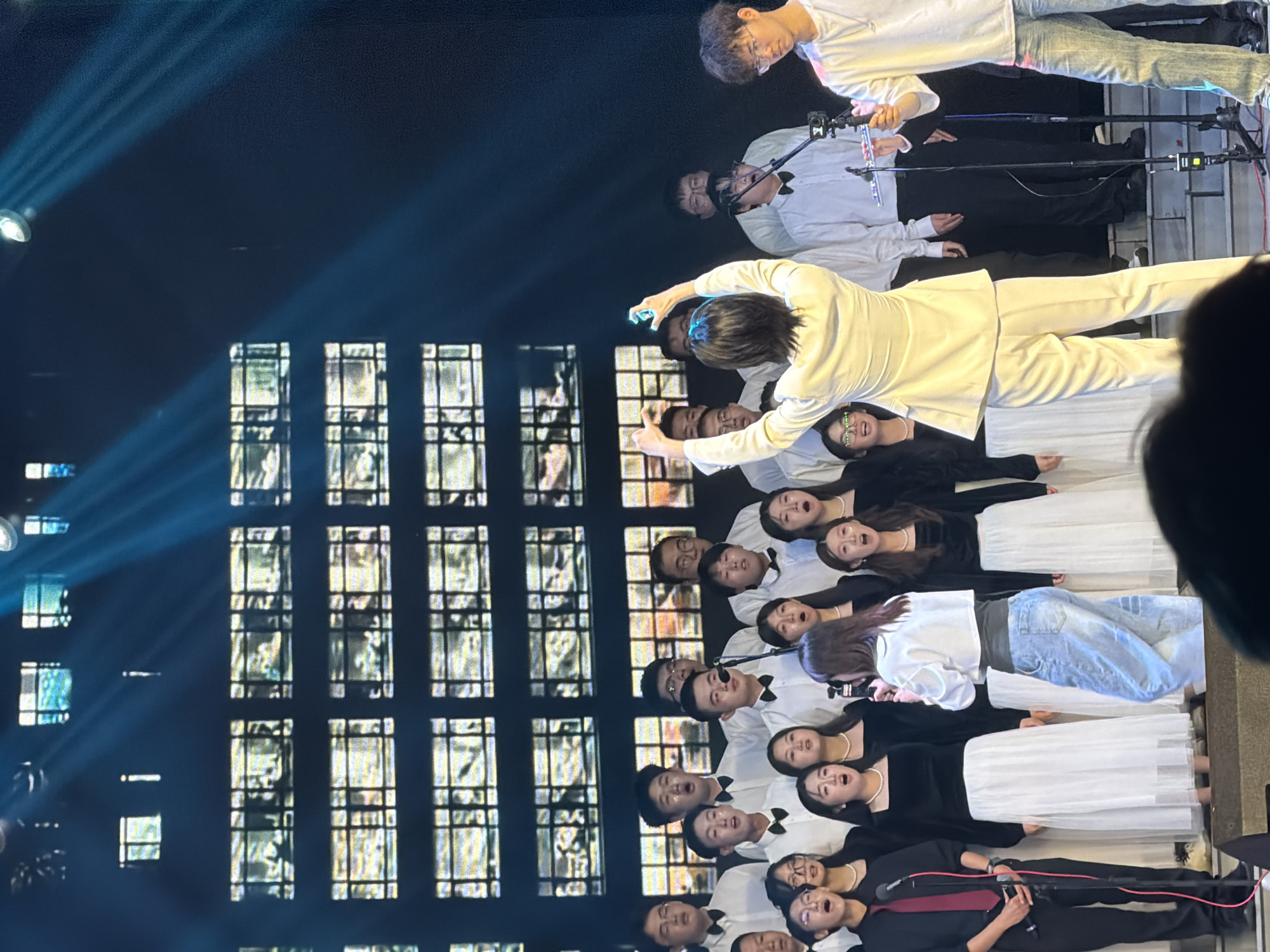 The image depicts an indoor musical performance, likely a choir concert or vocal ensemble, taking place in Guta, China. The setting is a stage or performance area with theatrical lighting.
In the foreground, a central figure dressed in a white suit stands prominently, appearing to be a lead vocalist or conductor, with an arm extended in a gestural pose. Directly behind and to their side, several male performers are visible, uniformly dressed in white shirts and black bow ties, some holding microphones on stands.
Further back, a large choir is arranged, primarily composed of women. They are dressed in black tops and long white skirts. Many members have their mouths open in song, indicating active vocalization. Among the choir, a person in a white top and blue jeans is also visible, holding a microphone, possibly a soloist or additional performer. Another individual in a black top, red tie, and black pants is also seen with a microphone, suggesting diverse roles within the performance.
The background consists of a dark wall with a grid pattern of illuminated rectangles, resembling stylized windows or a modern architectural feature, which serves as a backdrop for the performance. Strong beams of blue-tinted light from overhead spotlights cut across the dark upper part of the scene, creating a dramatic atmosphere. The scene is well-lit specifically in the performance area, while the surroundings remain dark, suggesting an evening event or a dedicated performance space. The participants appear focused and engaged in the musical presentation. No visible text is present in the image.