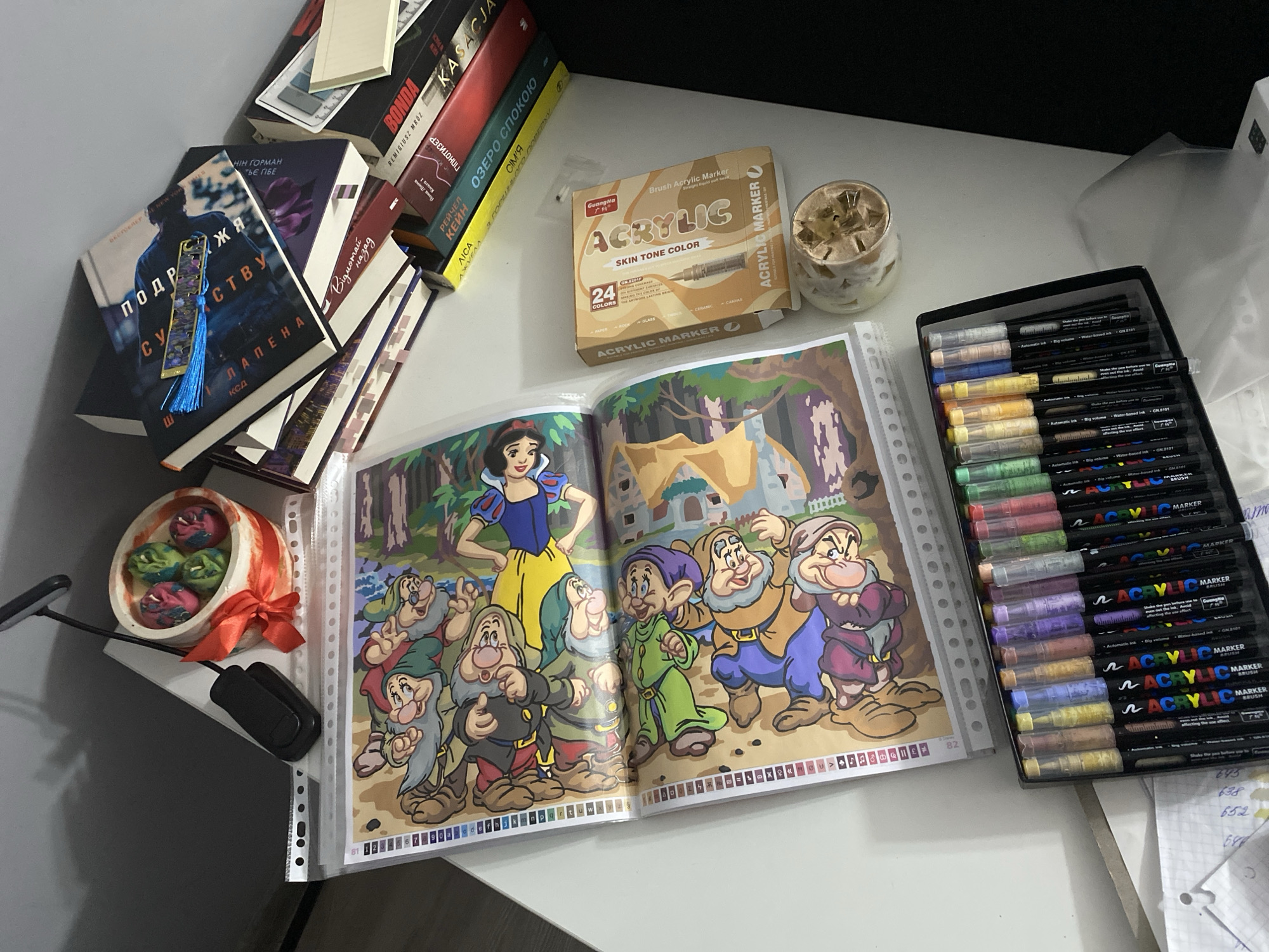 The image displays a desk with various art supplies and books. In the foreground, a box of acrylic markers is laid out, featuring a range of colors. To the right, a box of "ACRYLIC MARKER" in "SKIN TONE COLOR" is visible, alongside a glass containing a light-colored beverage with ice and what appears to be crushed fruit or cereal. Behind the markers, an open binder displays a coloring page featuring characters from Disney's Snow White and the Seven Dwarfs. Further back, a stack of books is arranged, with titles partially visible in what appears to be Cyrillic script. A small decorative item with a red ribbon and colorful items inside a cylindrical container sits near a black object, possibly a device accessory. The scene is indoors, with the lighting suggesting it is either natural light from a window or artificial lighting. There are no people in the image. The image does not contain any information that could be used to determine its location in Łódź, Poland.