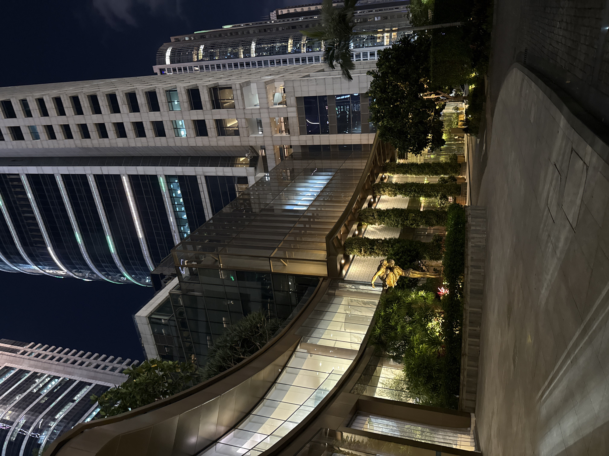 View of Guangzhou city in Tianhe District, China at night. The image captures a ground-level perspective looking up at modern architecture. Several buildings are visible, including one tall, slender skyscraper with a curved, illuminated facade. Another building features a more traditional architectural style with regularly spaced windows. A multi-level structure with glass walls and a curved design is situated closer to the viewer, containing plants, shrubs, and a bronze-colored statue of two figures. A sidewalk runs along the right edge of the image, lit by streetlights. The night sky is visible in the background.