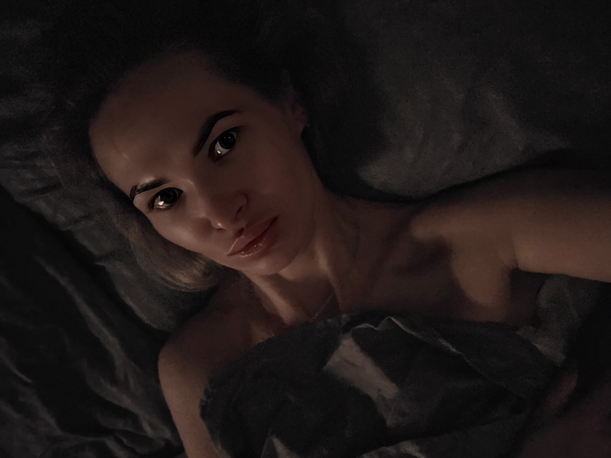 A selfie-style, medium-shot photograph depicts a woman lying down, seemingly in a bed. Her head is at the top left, and she is looking directly at the camera with a neutral expression. Her shoulders and upper chest are visible; she appears to be partially covered by a dark grey blanket. The lighting is dim, creating heavy shadows. The background seems to be a dark pillow or the head of the bed. No identifying information is present that would allow determining the location in the city of Chaikovsky, Russia.