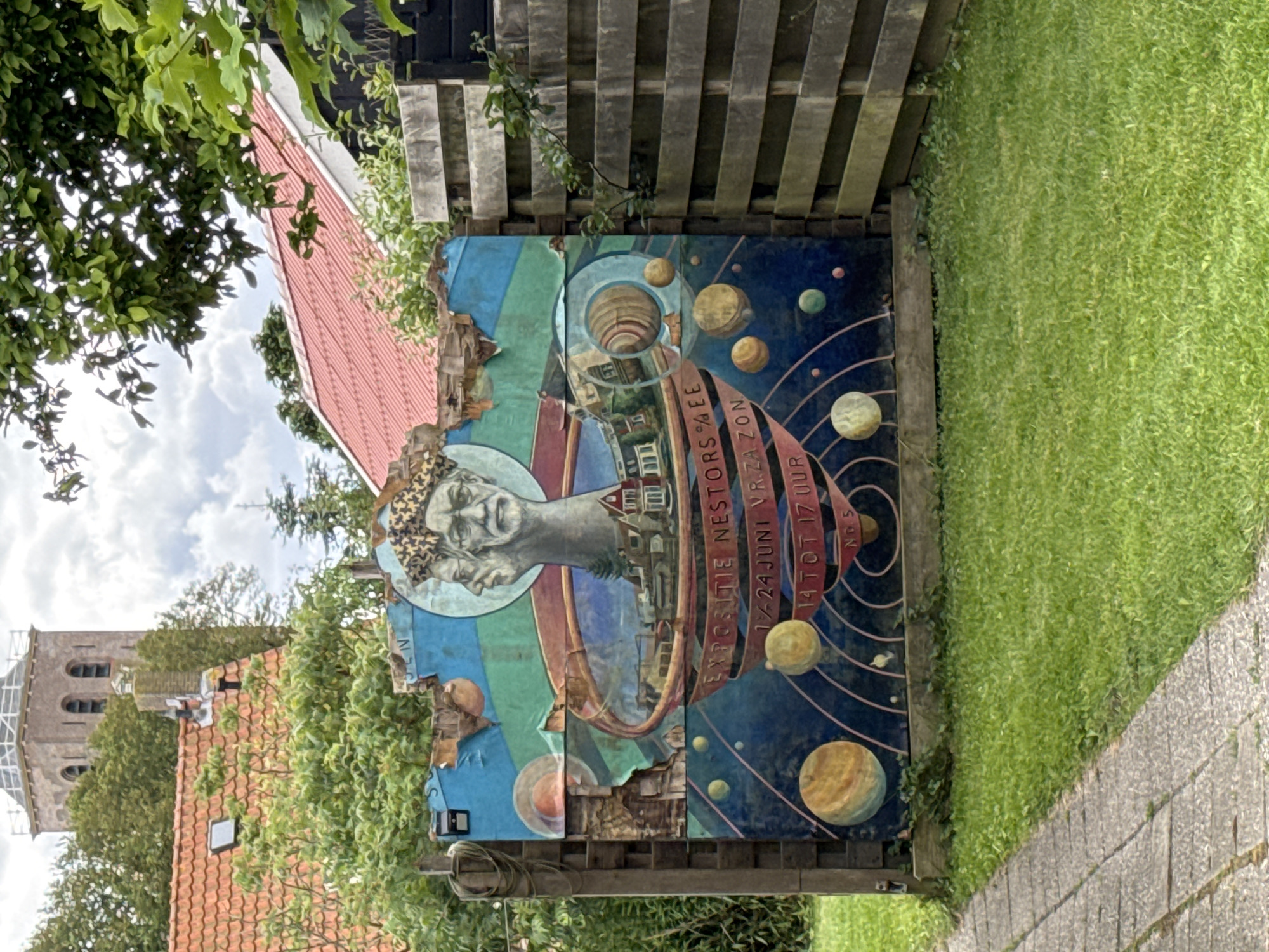 The image depicts a vibrant, weathered mural on a wooden structure in what appears to be a residential or garden setting. The mural features a stylized depiction of a head with two faces, a crown, and a celestial theme with planets and orbits. Text on the mural reads "EXPOSITIE NESTOR" and a date range "14-24 JUNI VRZ. AZON" and "14 TOT 17 UUR", suggesting it is an advertisement for an exhibition. The mural is attached to a wooden fence, and a stone pathway leads through a grassy area in front of it. In the background, a church tower and red-tiled roofs of houses are visible, along with lush green trees and foliage. The sky is partly cloudy. The mural itself shows signs of age and damage, with some sections peeling and torn.