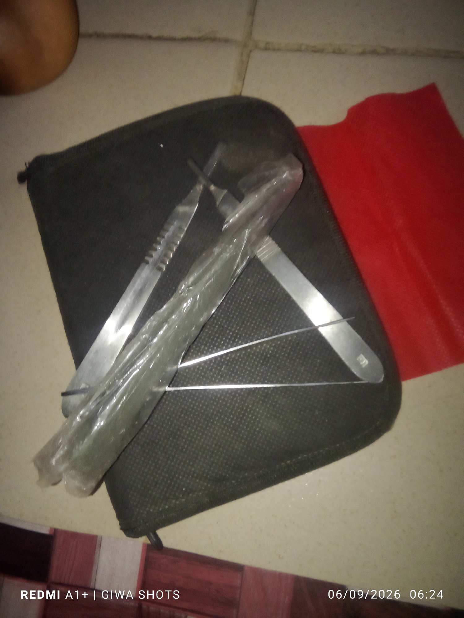 A high-angle medium shot captures a dark gray zippered pouch containing a set of metal instruments. These instruments include a scalpel with a serrated edge and another instrument with a smooth, flat blade. A set of fine metal probes is also visible, some of which are wrapped in clear plastic. A bright red piece of material is partially visible to the right of the pouch. The scene appears to be on a tiled floor with a light beige color. The bottom of the image displays a timestamp and camera model: "06/09/2026 06:24 REDMI A1+ | GIWA SHOTS".