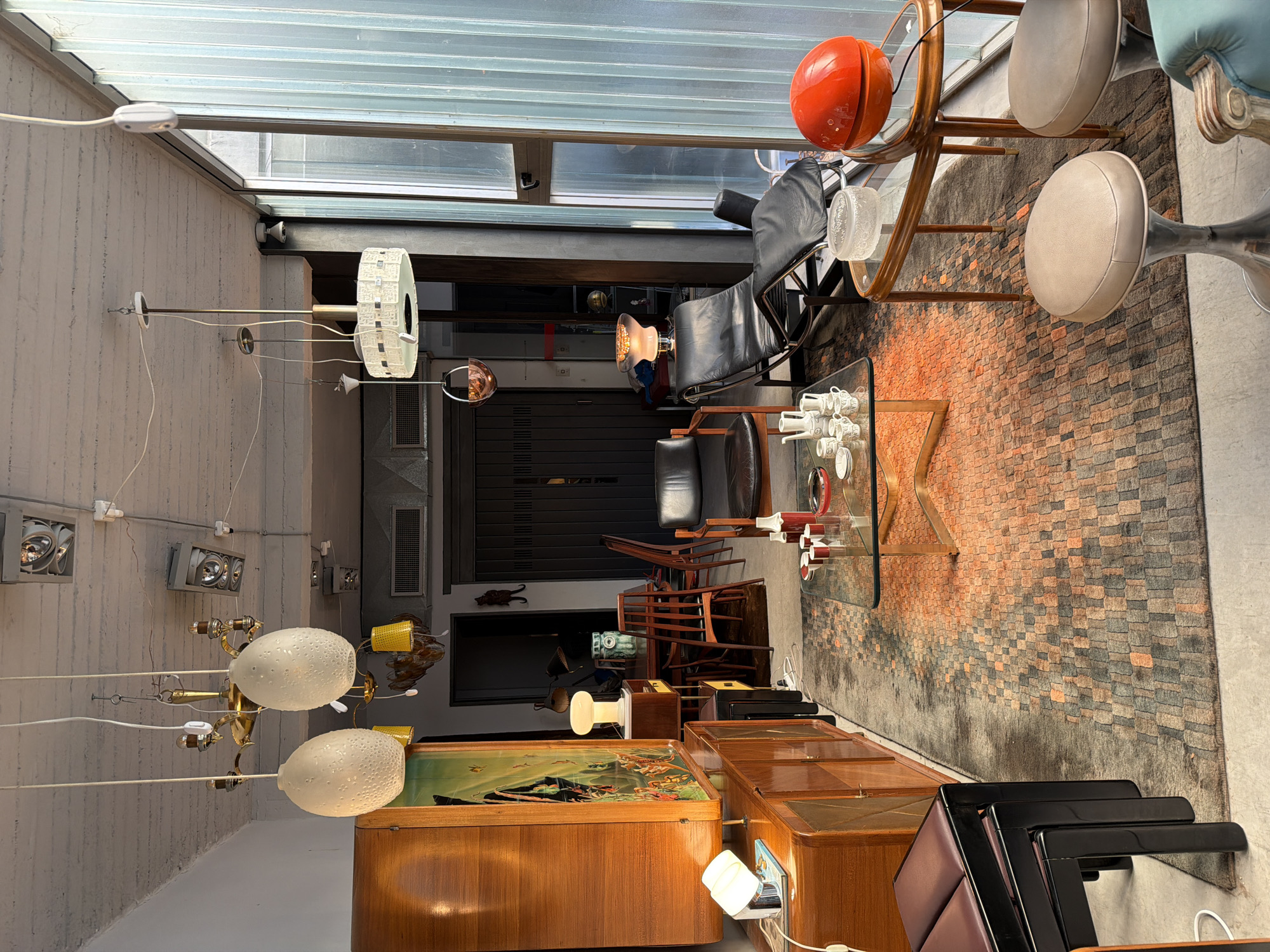 This image captures a bright, organized indoor space, likely a vintage furniture and lighting showroom or a curated living area in Buenos Aires, Argentina. The setting features concrete walls, industrial-style metal beams, and a large translucent corrugated glass ceiling that bathes the room in natural daylight.
The space is densely populated with a diverse collection of mid-century modern and vintage items. Prominently displayed are several polished wooden cabinets and sideboards, some with green tops. One cabinet showcases a painting depicting classical figures in a landscape. A variety of distinct light fixtures illuminate the scene, including two large, textured, egg-shaped pendant lights, a spherical copper-toned pendant, a tall floor lamp with a wide, flat circular shade, and several wall-mounted and track lights. A small, white, mushroom-shaped table lamp adds a whimsical touch.
Other furniture includes a pair of black leather lounge chairs, a glass-top coffee table with a wooden frame, and several beige upholstered stools with polished metal bases. A large, patterned rug featuring a mix of orange, grey, and black brick-like shapes covers the floor. On the coffee table, a bright orange spherical decorative object sits alongside a stack of white ceramic cups and saucers. In the background, stacked wooden chairs, a dark roll-down shutter, and various other decorative objects like a cat figurine and a globe are visible.
No people are present, and there is no specific activity taking place, suggesting the scene is arranged for display or presentation. The bright illumination indicates it is daytime. The overall ambiance is one of curated design, blending industrial architectural elements with a rich assortment of retro and stylish furnishings. No visible text is present in the image.