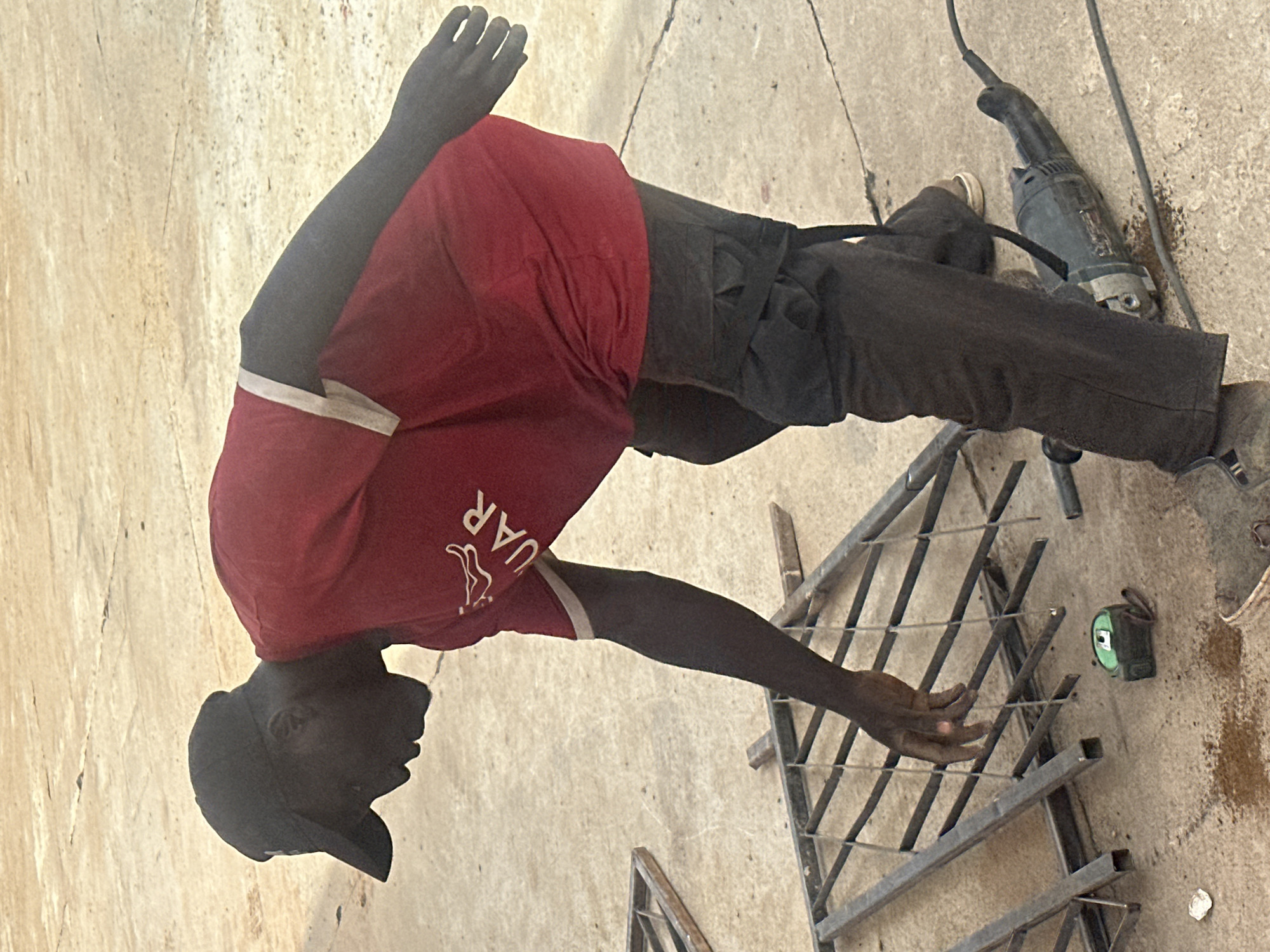 A young Black man wearing a red t-shirt with white trim and a black baseball cap is working on a metal structure outdoors. He is in profile, facing left, with his right arm extended towards the metal bars. His t-shirt has white lettering and a graphic of a leaping animal. He is wearing dark pants and appears to be barefoot or wearing very light-colored shoes.
In the background, a black angle grinder with a cord is visible on the ground, along with some metal shavings and dirt. A green measuring tape is also on the ground near the metal structure. The ground appears to be made of concrete or a similar rough surface. The lighting suggests it is daytime, likely sunny given the shadows. The overall scene depicts a person engaged in manual labor, possibly welding or assembling metalwork. The location is identified as Barakin Lerawi, Nigeria. There is no clear indication of weather or specific emotions.