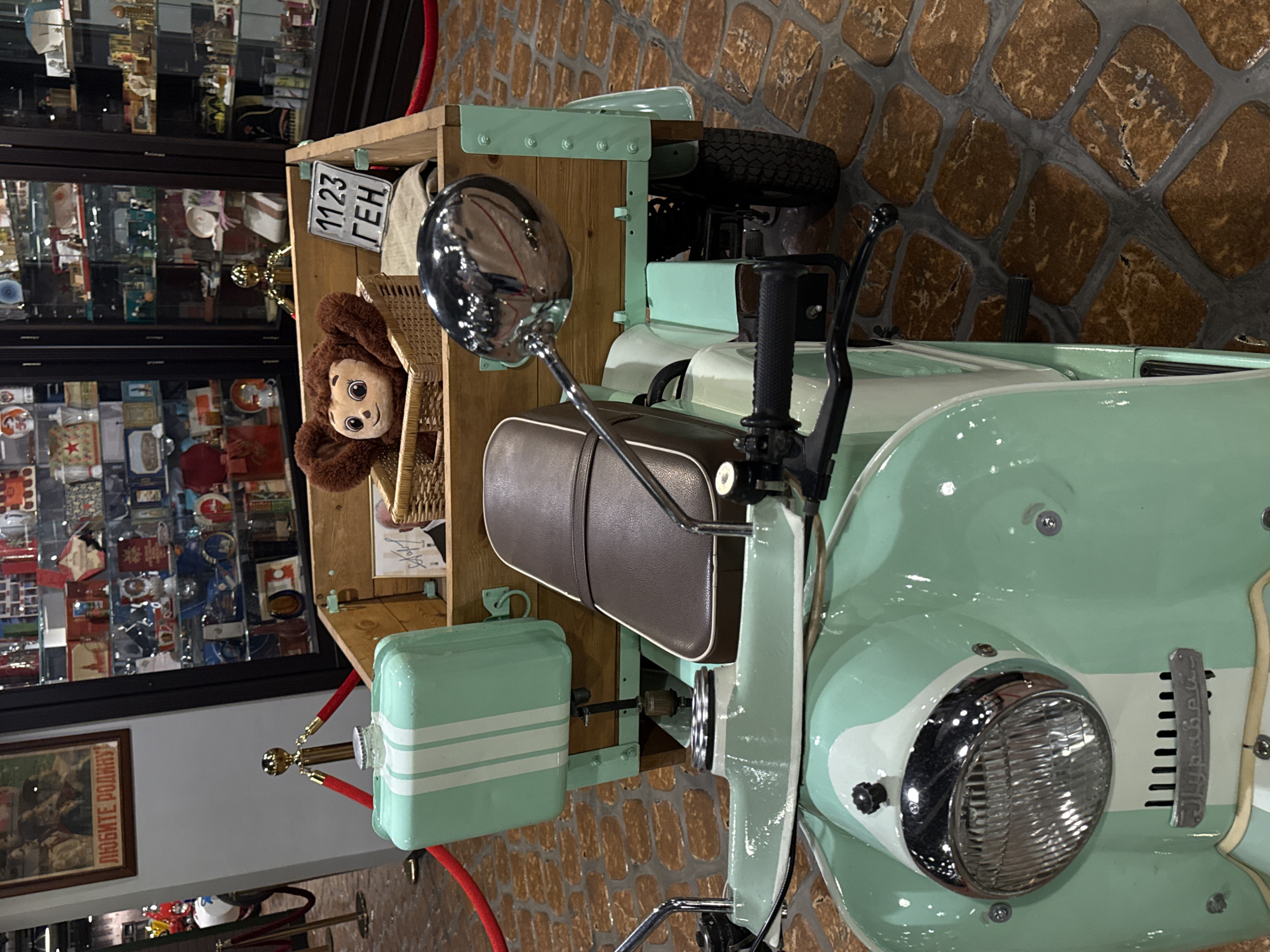 This is a close-up, eye-level shot of a vintage mint green scooter with a wooden cargo box attached. The scooter has a brown leather seat and a chrome headlight. The cargo box contains a wicker basket with a Cheburashka plush toy and a small license plate with Cyrillic lettering that reads "1123 ГЕН". In the background, glass display cases filled with various items are visible, suggesting a museum or exhibition setting, possibly the Museum of Toys in Arkhangelskoye, Russia, given the style of the exhibits. The scooter is parked on a patterned floor made of reddish-brown stone-like tiles. Red velvet ropes on gold stanchions frame the scene, indicating a restricted area.