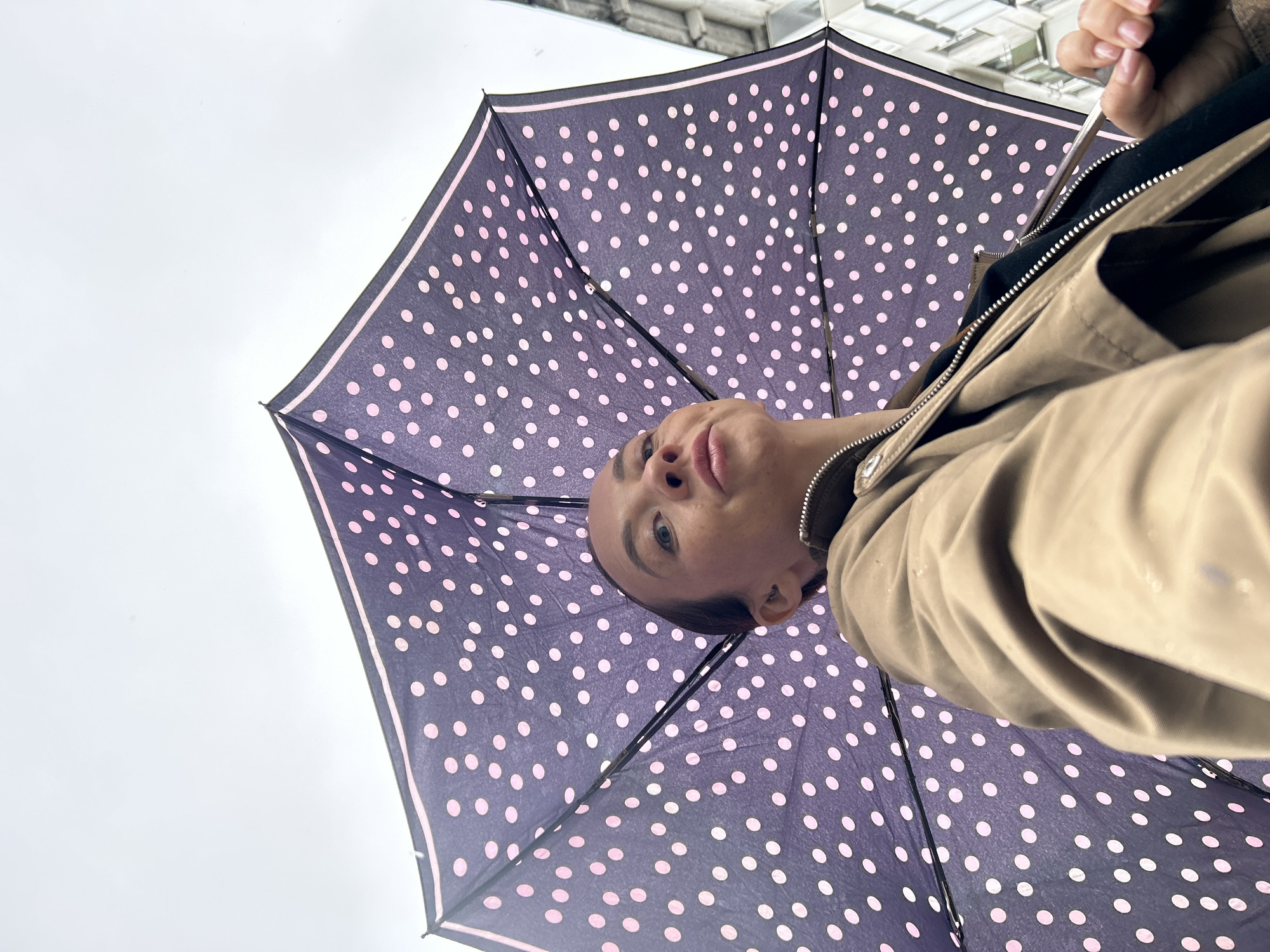 A low-angle shot depicts a person holding a polka-dot umbrella. The umbrella is dark blue with evenly spaced light pink polka dots, and a thin light pink trim along the edge. The person's face is visible from below, showing their head, chin, nose, and eyes. They are wearing a light brown jacket. In the background, a gray sky is visible. Based on the request, the location is Chaikovsky, Russia, though no landmarks or other specific details in the image confirm this.