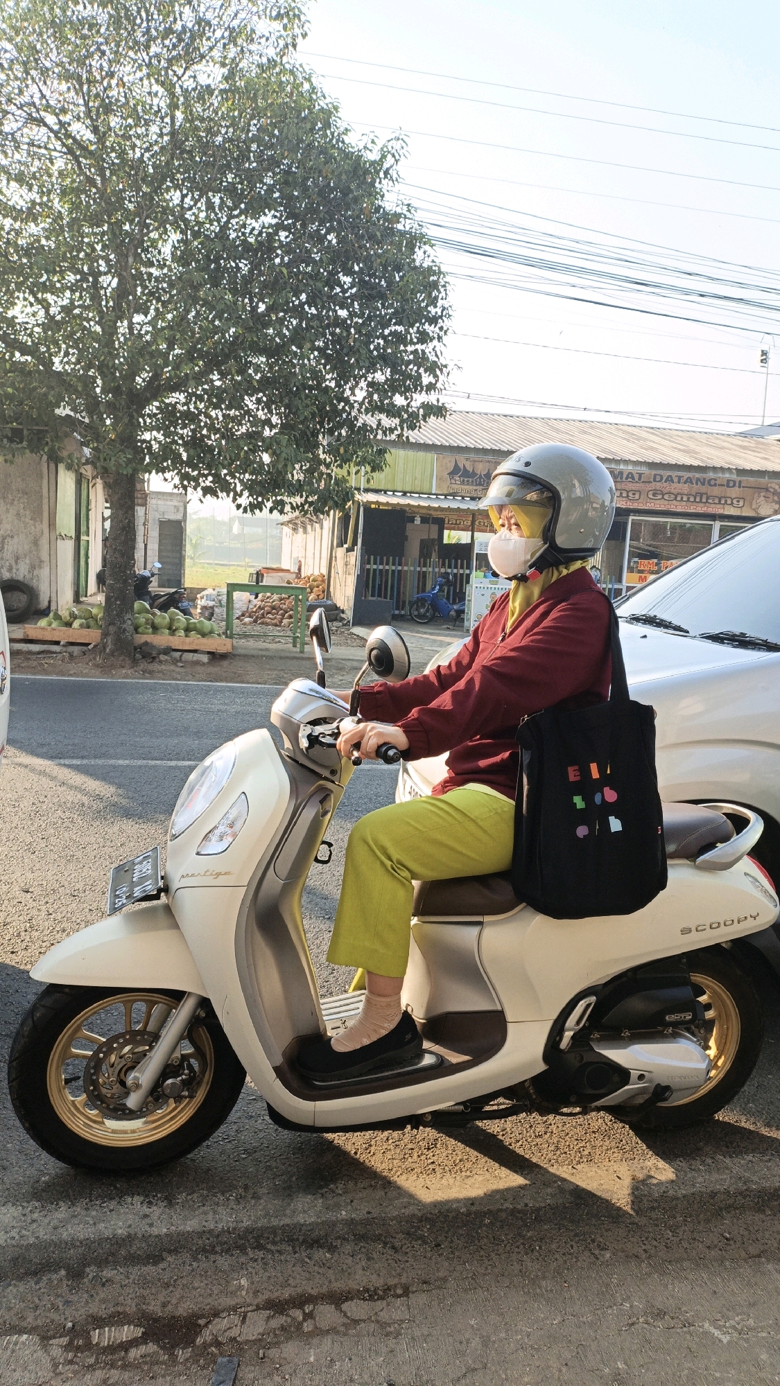 In Kuwasen, Indonesia, a person wearing a light grey helmet with a yellow visor, a white face mask, a maroon long-sleeved shirt, and bright green pants is seated on a white scooter. They are holding onto the handlebars and appear to be stopped on the side of a paved road. A black tote bag with colorful geometric shapes is hanging from their shoulder. The scooter has a license plate on the front and the word "Scoopy" is visible on the rear. In the background, there are several buildings, including one with a sign that reads "SELAMAT DATANG DI" followed by Indonesian text. Piles of green coconuts are displayed on wooden pallets in front of some of the buildings. A car is parked next to the scooter. The scene is illuminated by bright sunlight, casting shadows on the road.