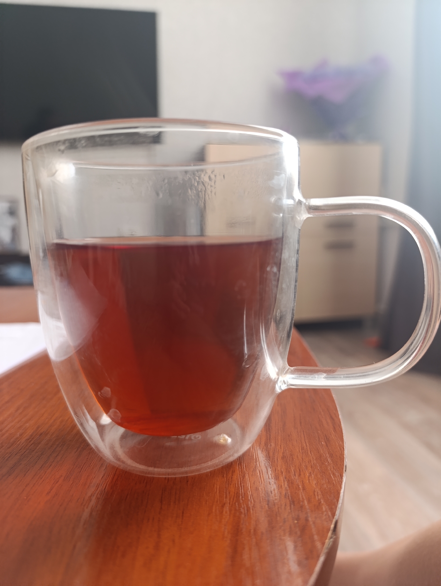 A close-up shot focuses on a clear, double-walled glass mug filled with a dark amber liquid, likely tea. The mug is positioned on a polished wooden table, its surface reflecting the warm tones of the wood. The background is softly blurred, hinting at an indoor setting. To the left, a dark rectangular object, possibly a television screen, is visible. In the mid-ground to the right, a cabinet with light-colored doors and drawers can be seen, adorned with a vase containing purple flowers. The lighting suggests it's daytime, with sunlight casting a gentle glow and creating highlights on the mug and table. The overall atmosphere is calm and domestic. There is no visible text in the image.