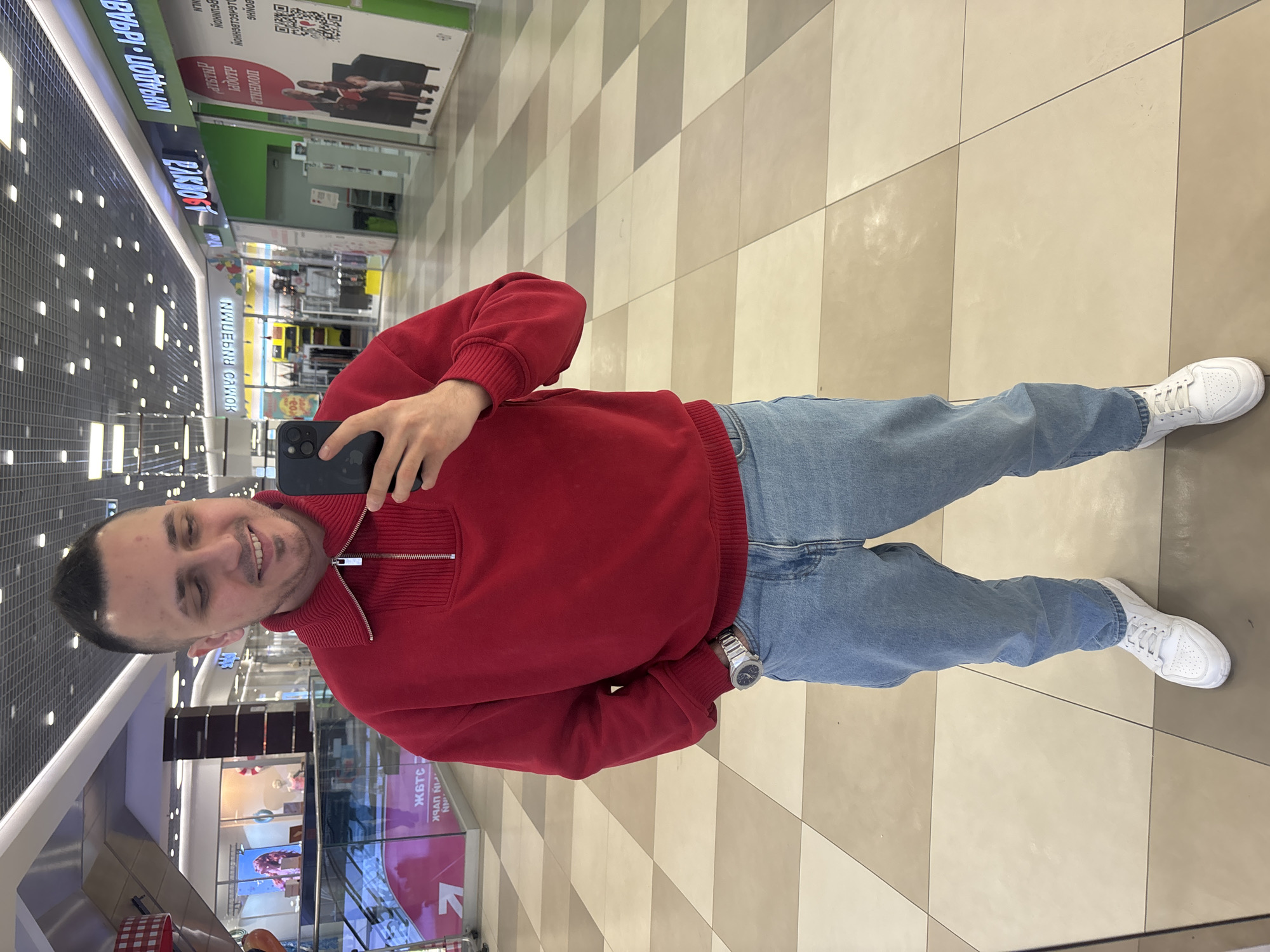 The image shows a man taking a selfie in what appears to be a shopping mall. He is wearing a red sweater, blue jeans, and white sneakers. He is holding a smartphone in his right hand and smiling at the camera. The mall has a checkered floor and various storefronts visible in the background. The mall is possibly located in Saint Petersburg, Russia, as suggested by the Cyrillic text visible in the signs.