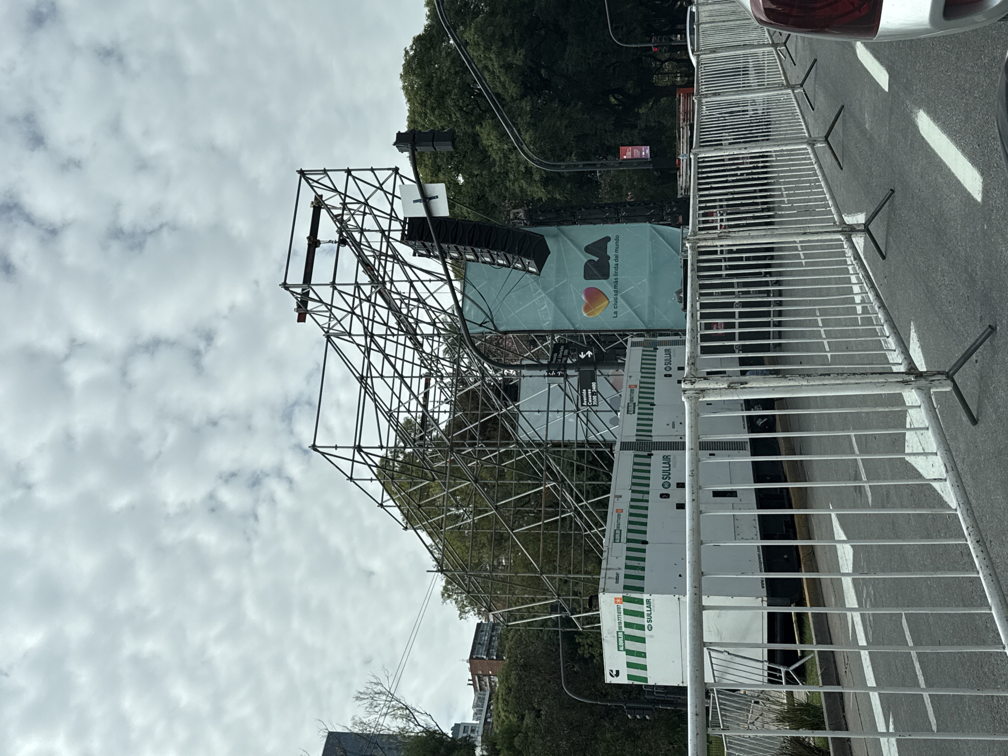 The image shows a street scene in Buenos Aires, Argentina. A large metal scaffolding structure is erected near the road, with a banner partially visible that reads "La ciudad mas linda del mundo" (The most beautiful city in the world) with a heart-shaped logo. Several Sulair generators are located next to the structure. In the foreground, there are metal crowd control barriers. The street has white lane markings, and a car is partially visible on the right side. In the background, there are trees and buildings, along with a traffic light. The sky is overcast. The street number is Avenida Casares 3500-3400.
