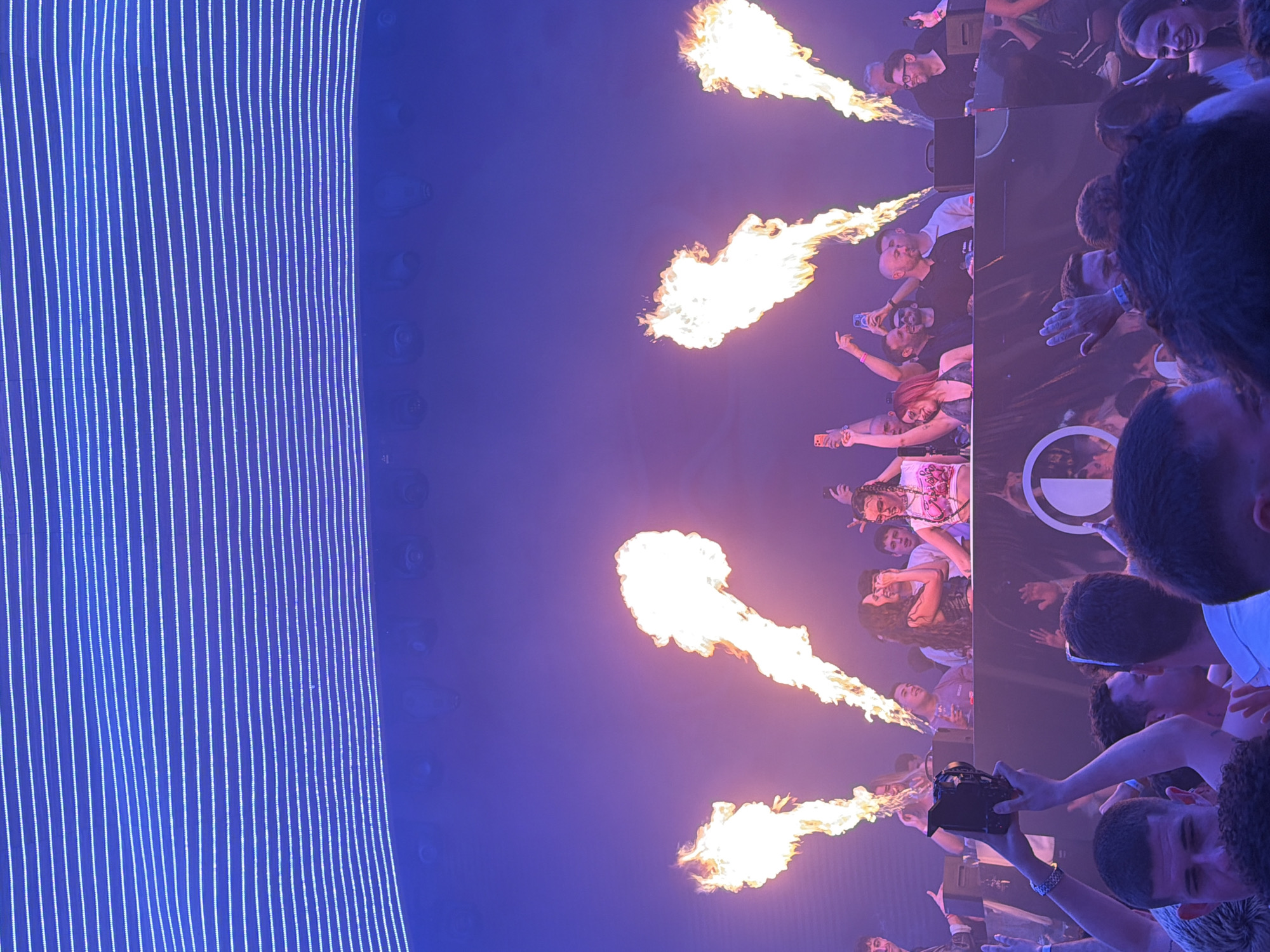 The image depicts a concert or event, possibly in Dénia, Spain, given the prompt context. Four bursts of flames are projected upwards in a line. A crowd of people is visible, mostly with their backs or sides facing the camera, suggesting they are watching a performance. Some are holding up phones to record or take photos. A light structure with vertical blue lines appears on the left. The overall environment is dark, illuminated by stage lighting and the flames. The scene suggests a lively and energetic atmosphere with special effects.