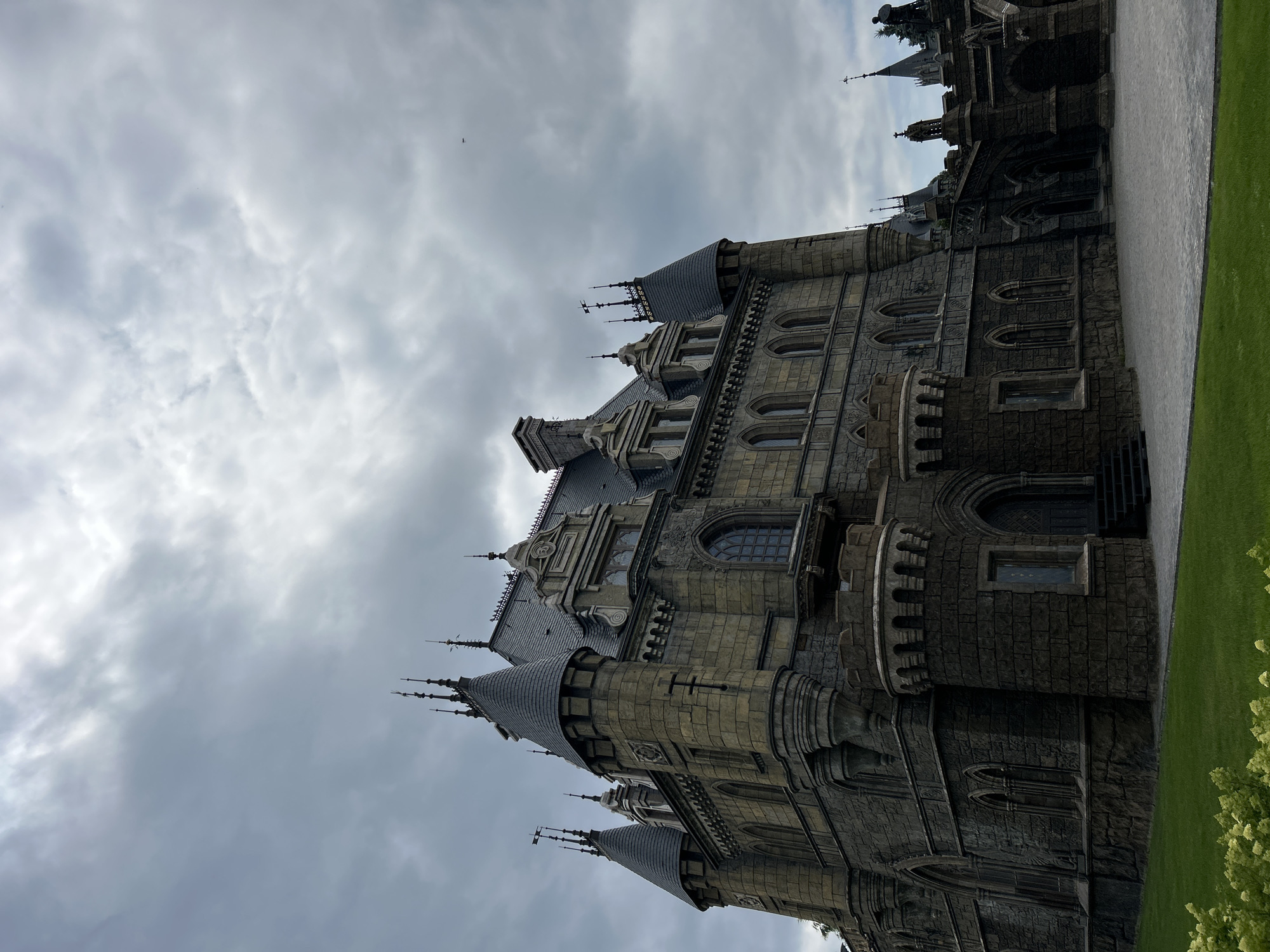 The image shows a large, gothic-style castle, likely the Von Skall and Von Kremer Castle, located in Khryashchevka, Russia. The castle is constructed from dark stone, with multiple towers, spires, and intricate architectural details, including arched windows and decorative stonework. The building is imposing and appears to be in good condition. The sky above is overcast with dramatic, gray clouds, suggesting a somber or moody atmosphere. In the foreground, there is a manicured green lawn with some light-colored foliage. A paved courtyard area is visible in front of the castle. No people are present in the scene.