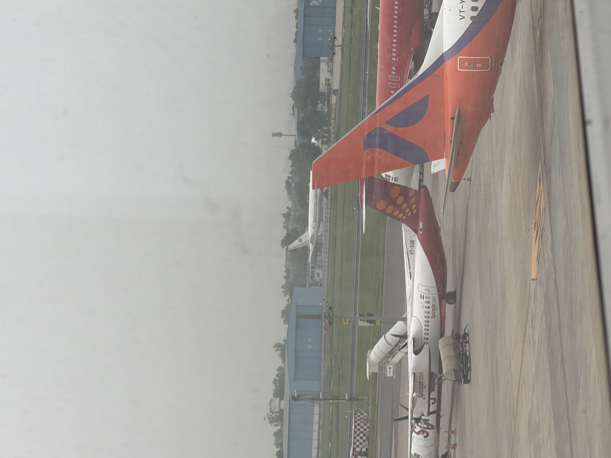 The image depicts an airport scene, likely at Indira Gandhi International Airport in New Delhi, India, due to the presence of aircraft with Indian registration markings. Several airplanes are visible on the tarmac. In the foreground, a SpiceJet turboprop aircraft is parked, with its propellers and the "SpiceJet.com" livery clearly visible. Adjacent to it, to the right, is the tail of a larger, red-colored airplane. Further in the background, another white airplane with a red tail section is parked, and beyond that, a larger white passenger jet is seen on the runway or taxiway. Blue hangars or airport buildings are situated in the far background, along with trees and a hazy sky, suggesting a possibly overcast or dusty day. The overall scene is static, showing parked aircraft and airport infrastructure.