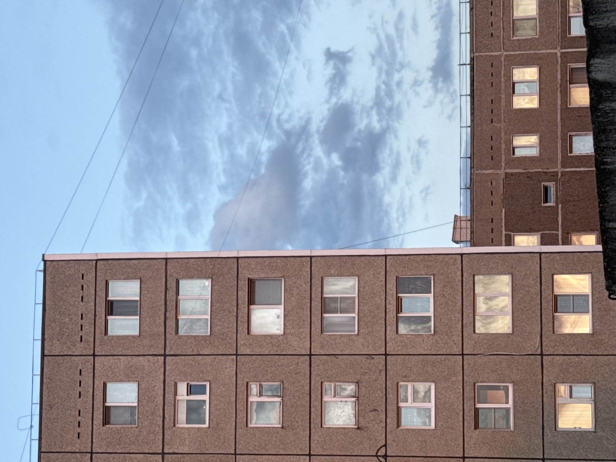 A low-angle shot depicts the facade of an apartment building in Chaikovsky, Russia. The building is constructed from concrete panels with square windows, some reflecting the cloudy sky above. A second building partially frames the shot on the right. Power lines cross the upper left corner of the frame. The sky is visible between the buildings and is pale blue with gray clouds. The overall impression is of a utilitarian architectural style common in post-Soviet urban environments.