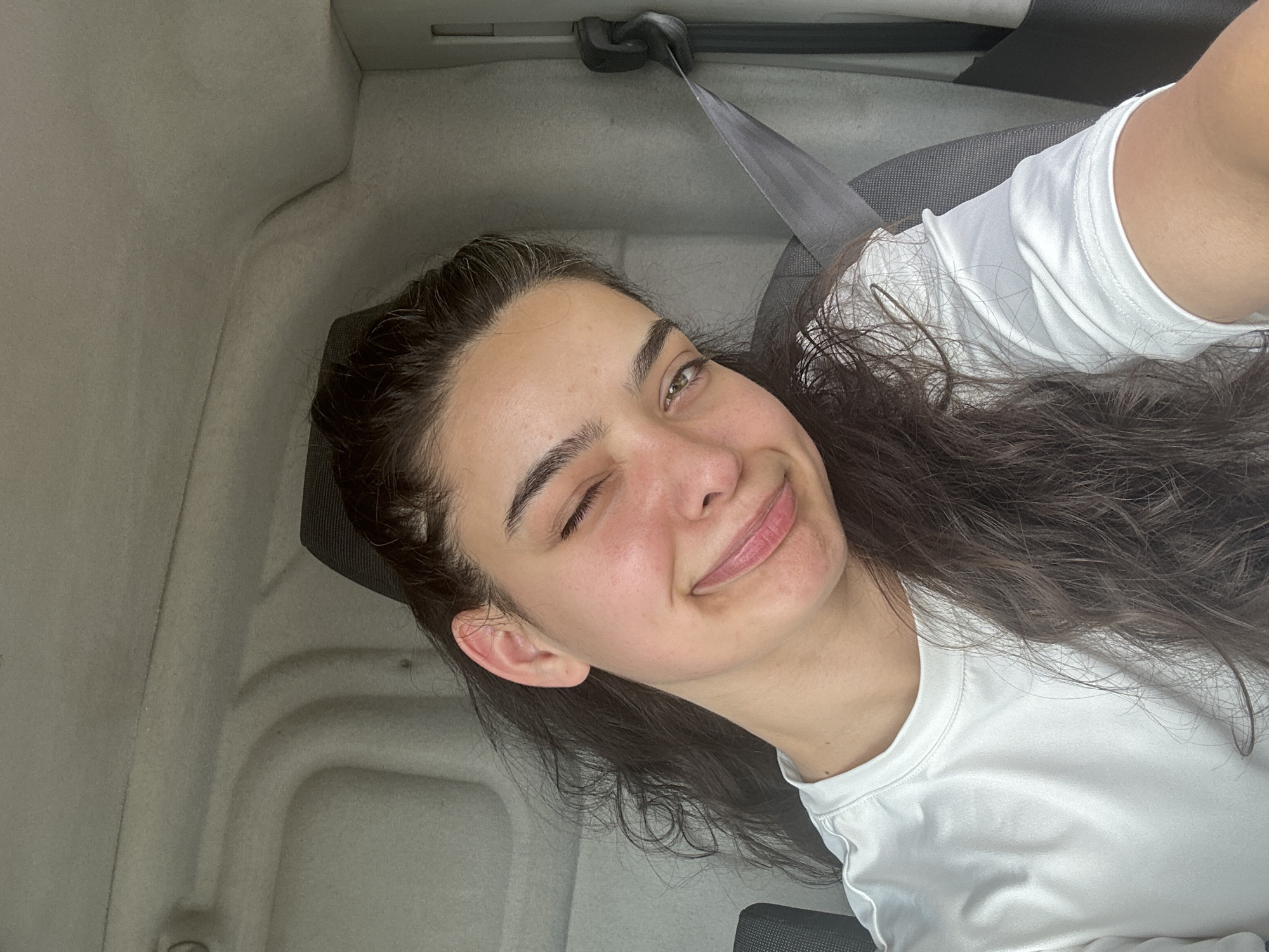 A young woman with long, dark, wavy hair and fair skin is sitting in the passenger seat of a vehicle, facing the camera. She is winking with her left eye and smiling with her lips slightly parted, revealing her teeth. Her right eye is open, and she has dark eyebrows and light-colored irises. She is wearing a white t-shirt. A grey seatbelt is visible across her chest, and the interior of the vehicle, including the headrest and ceiling, is visible in the background. The scene appears to be inside a car in Malevo, Bulgaria.
