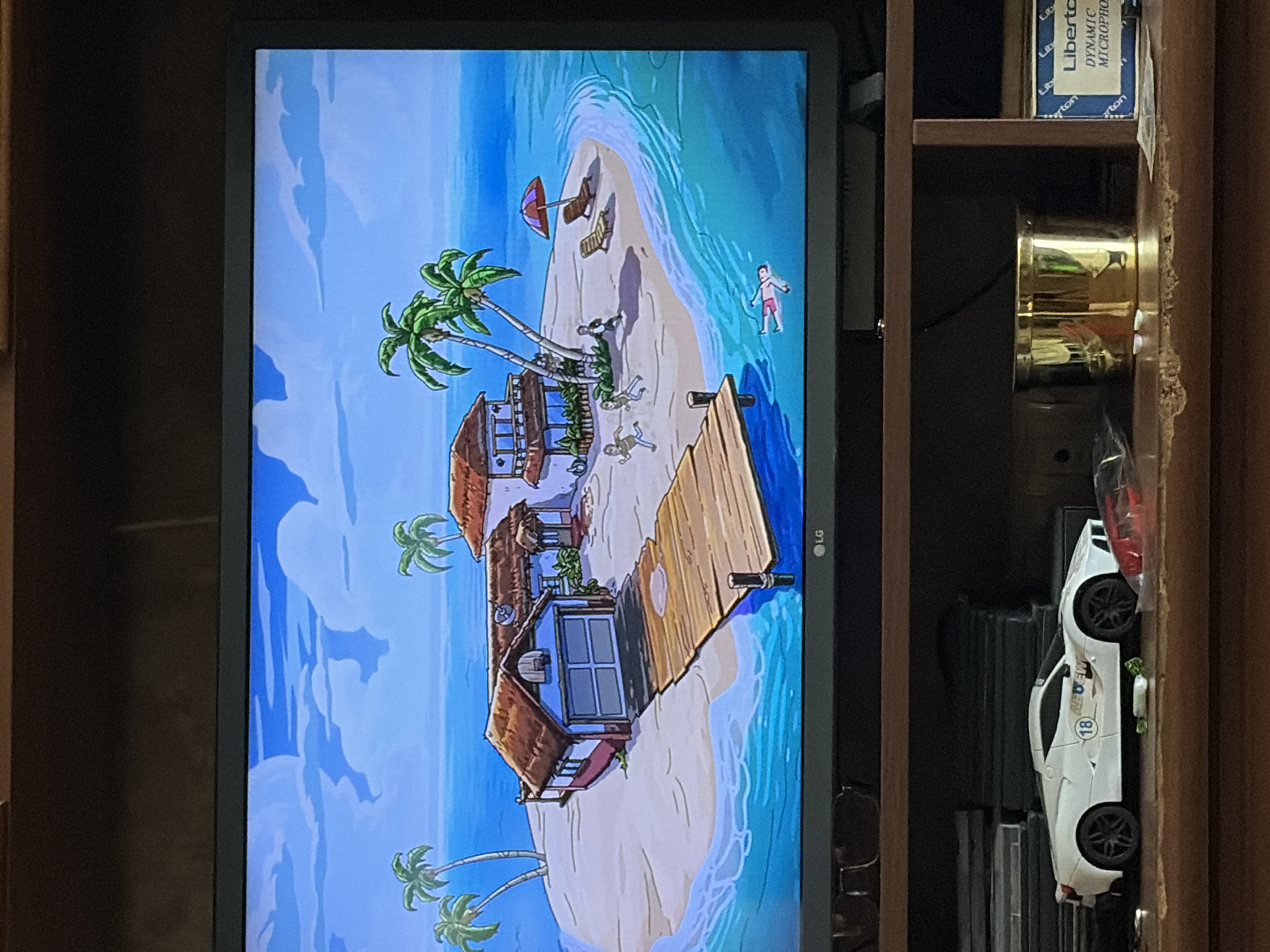 The image shows a television screen displaying an animated scene of a tropical island. The island features a beach, palm trees, two buildings, and a wooden dock. Several cartoon figures are depicted on the island, including some running and one person floating in the water. Below the television, on a wooden shelf, a white toy sports car is visible, along with other objects that are not clearly identifiable. The scene appears to be a screenshot from a video game or an animated show.