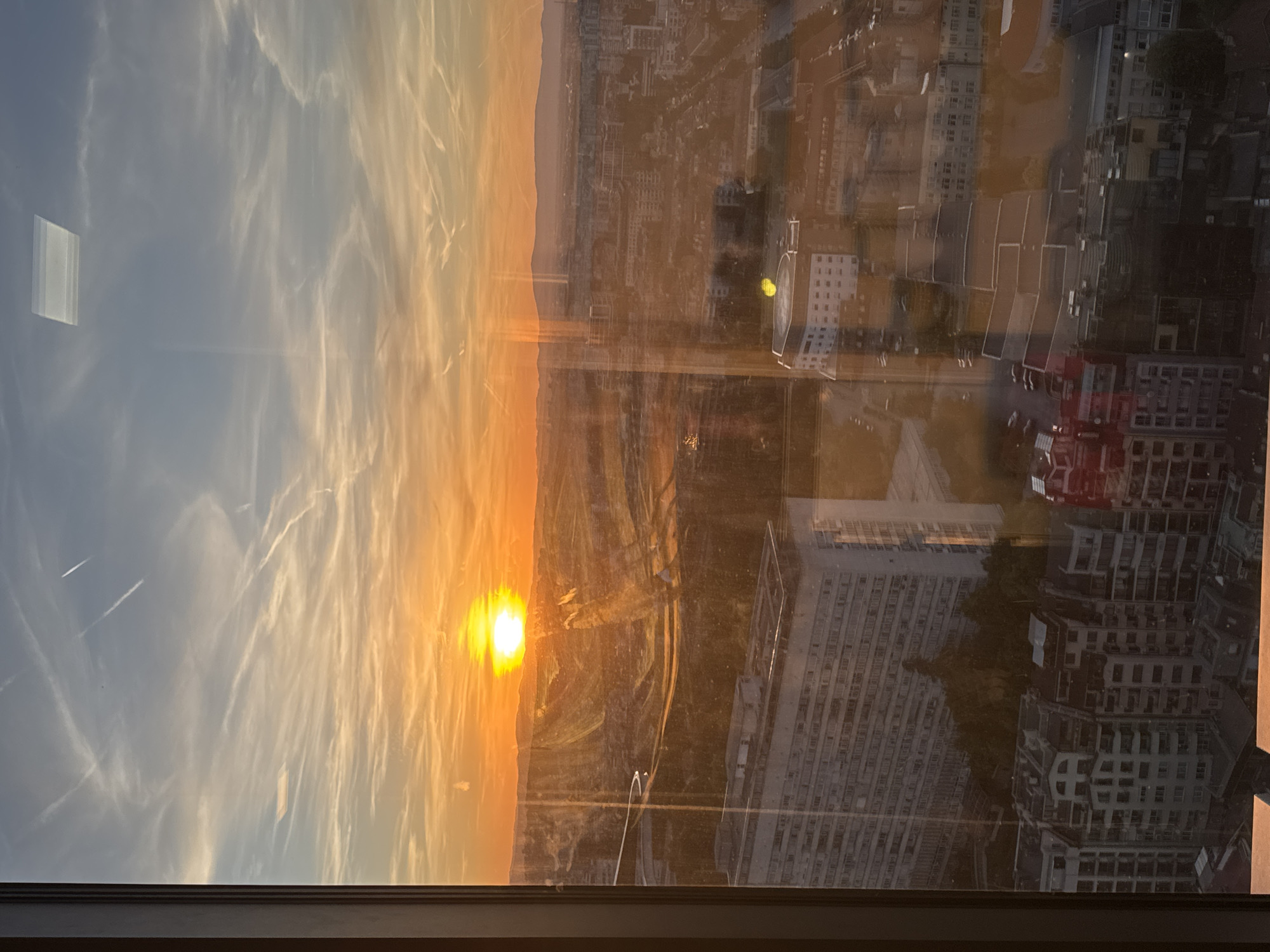 The image shows a sunset over Sofia, Bulgaria, viewed through a window. The sky exhibits an orange glow near the horizon where the sun is setting and transitions to a light blue hue higher up, with wispy clouds. A mountainous landscape is visible on the horizon. The window reflects the interior of a building, showing a cityscape with numerous buildings of varying heights and designs. Some buildings appear to be residential, while others seem to be commercial or office structures. Cars are parked on the streets. The window frame is visible at the bottom of the image.