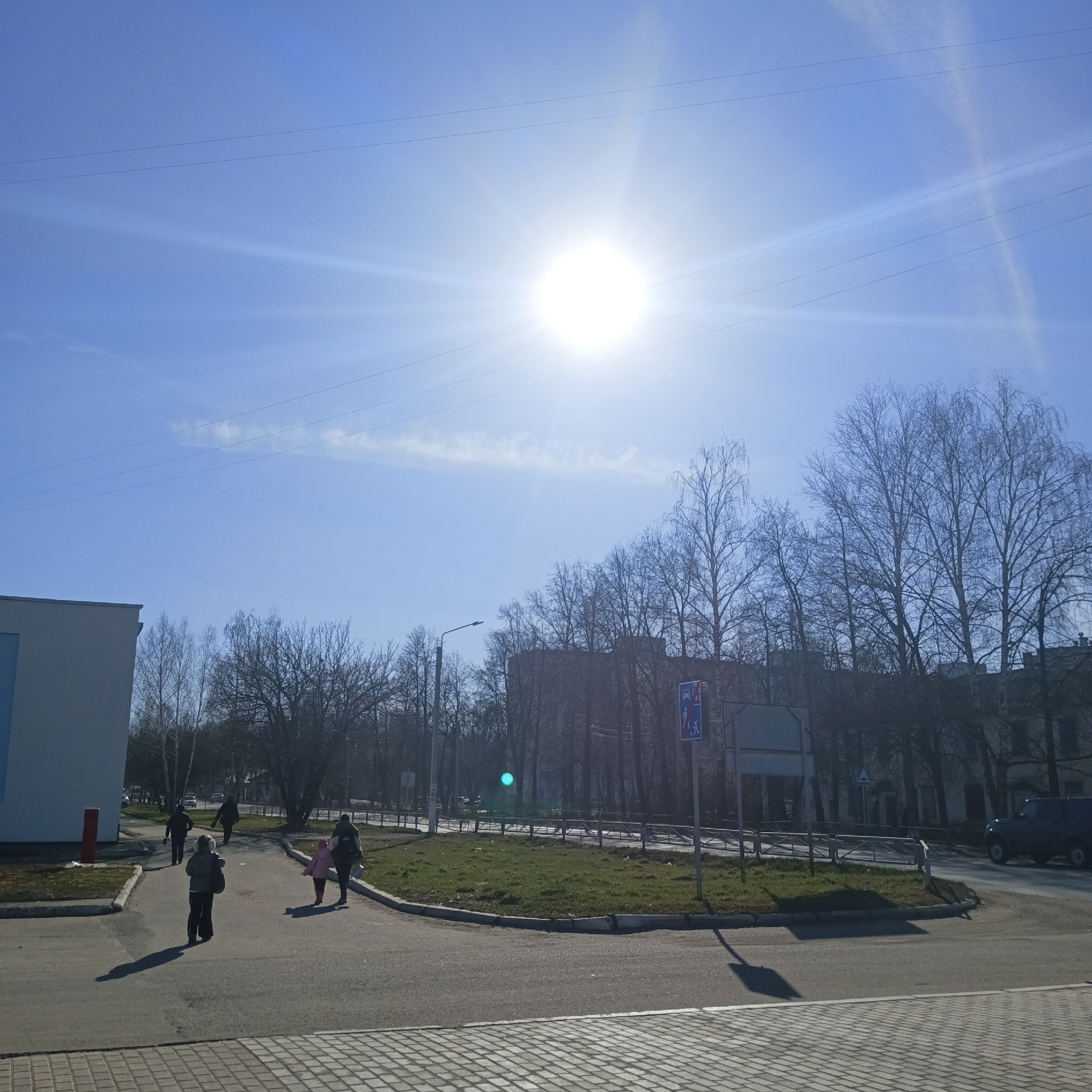 This image captures a bright, sunny day in an urban setting, likely in Chaikovsky, Russia, during late winter or early spring. The sky is a clear, vibrant blue, with the sun positioned high, creating a powerful lens flare and casting long, sharp shadows across the scene. Several thin power lines traverse the upper portion of the sky.
In the foreground and mid-ground, multiple individuals are walking on paved paths. A person in a dark jacket and trousers walks away from the viewer, with another woman and a small child in a pink jacket following nearby. Further back, two more people in dark attire are seen walking. The paths lead towards a central grassy median that shows patches of green, indicating the early stages of spring. This median is bordered by a low metal fence.
To the left, part of a light-colored building with a blue section is visible, along with a red post. The background is dominated by a dense array of deciduous trees, their branches bare, suggesting the season. Behind the trees, darker buildings can be faintly seen. A street lamp stands on the right side of the road.
A blue square traffic sign is prominently displayed on the right, featuring white pictograms. One section clearly shows a crossed-out pedestrian and bicycle symbol, likely indicating a restriction for these modes of transport. A dark SUV is partially visible on the road in the far right background. The overall scene depicts a calm, ordinary day with routine activity under bright sunlight.
