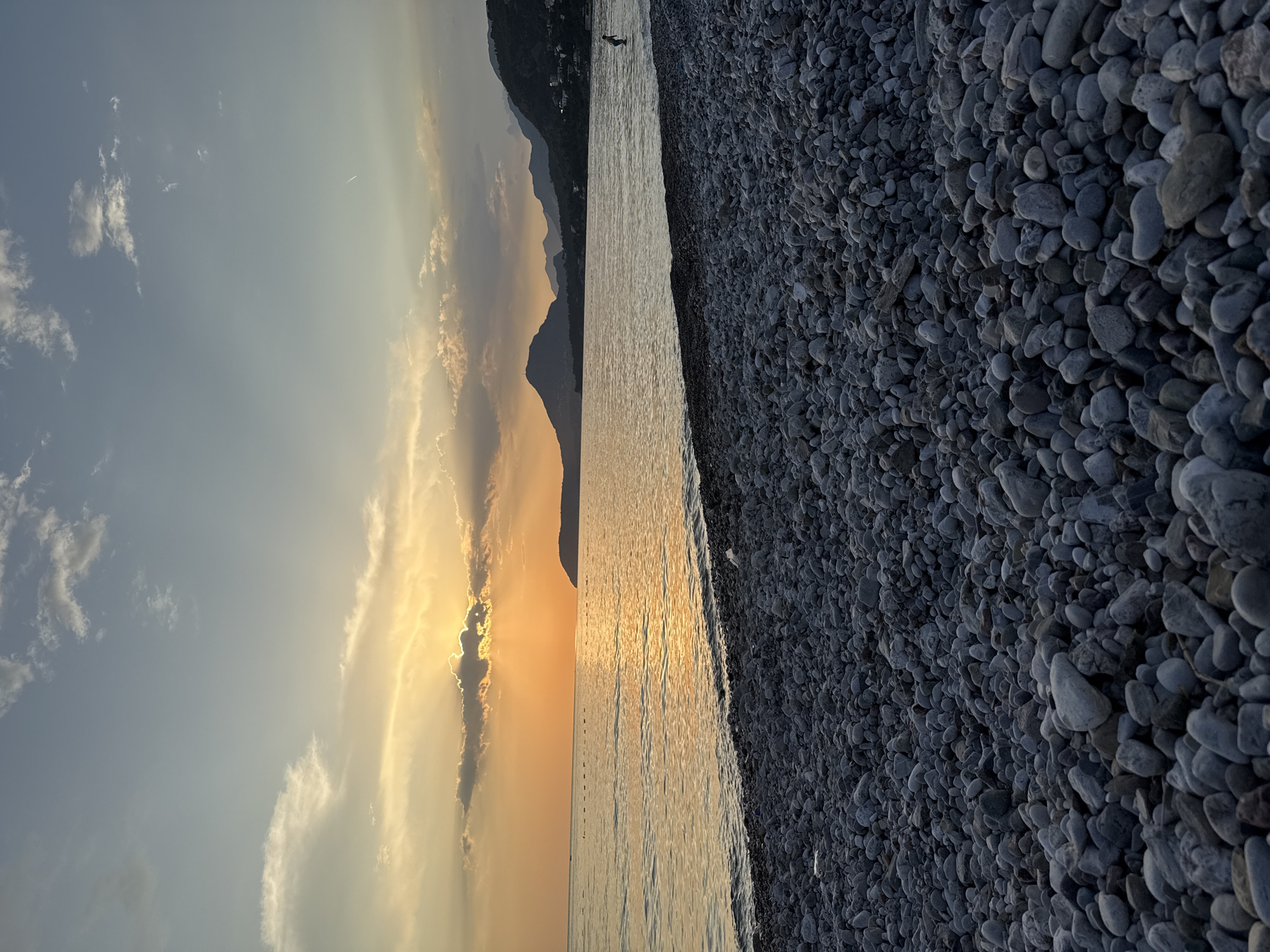 The image shows a landscape at sunset. The scene is divided vertically into three main sections: the sky, the sea, and the pebbly beach.
The sky section shows clouds around the setting sun. The light from the sun tints the clouds with golden and orange colors. The sea is a calm body of water reflecting the light from the sky.
The beach consists of smooth, grey pebbles. The pebbles extend into the foreground. On the horizon, mountains can be seen.
The location is likely in the city of Bar, Montenegro, given the pebbly beaches and mountain ranges in the area. There appears to be a person silhouetted on the beach in the distance.