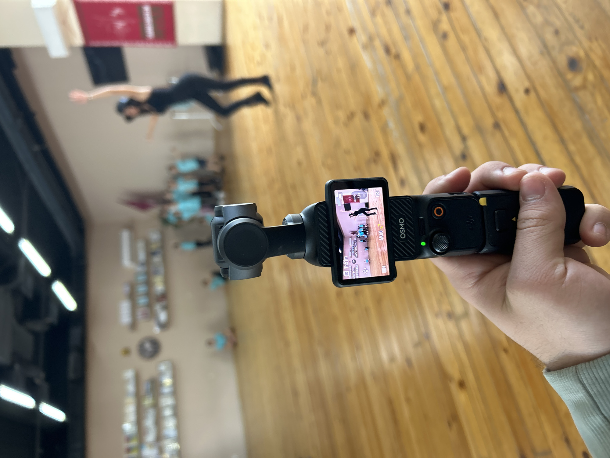 A person holds a DJI Osmo Pocket camera, capturing a scene in Varna, Bulgaria. The camera screen shows a recording of a dancer in a black outfit performing a mid-air pose. Several other people are visible in the background. The environment includes a wooden floor, bright overhead lights, and what appears to be a display case or shelves along a wall.