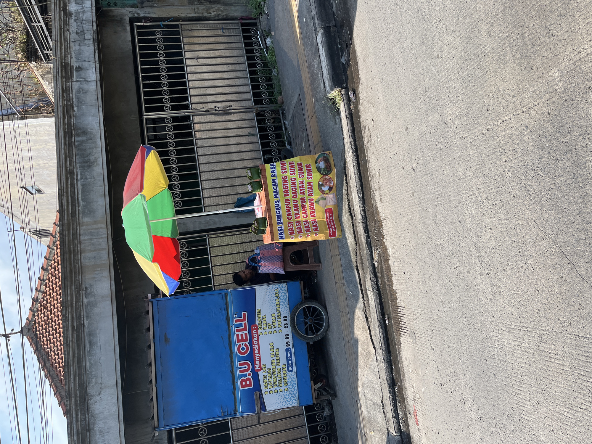 A mobile food cart, adorned with a multi-colored umbrella, is situated on a sidewalk in Tulangan, Indonesia. The cart, labeled "B.U CELL," displays a menu of items and services, including "GANTI LCD," "BATERAI," "TEMPERED GLASS," "SILIKON CASING," "VOUCHER," "CHARGER," "KABEL," "HF," and "PULSA/DANA.DLL." The cart is open from 09:00 to 23:00. Adjacent to the "B.U CELL" cart is another stall with a sign advertising "NASI BUNGKUS MACAM RASA" (Indonesian for "Rice Wraps of Various Flavors"). The menu on this stall lists "NASI CAMPUR DAGING SUWI," "NASI KRAWU DAGING SUWI," "NASI CAMPUR AYAM SUWI," and "NASI KRAWU AYAM SUWI." A person, presumably the vendor, is seated behind the "B.U CELL" cart, tending to a blue and pink woven bag. The background features a building with a tiled roof and a metal gate. The ground is paved with concrete and asphalt, with some debris and water visible on the roadside.