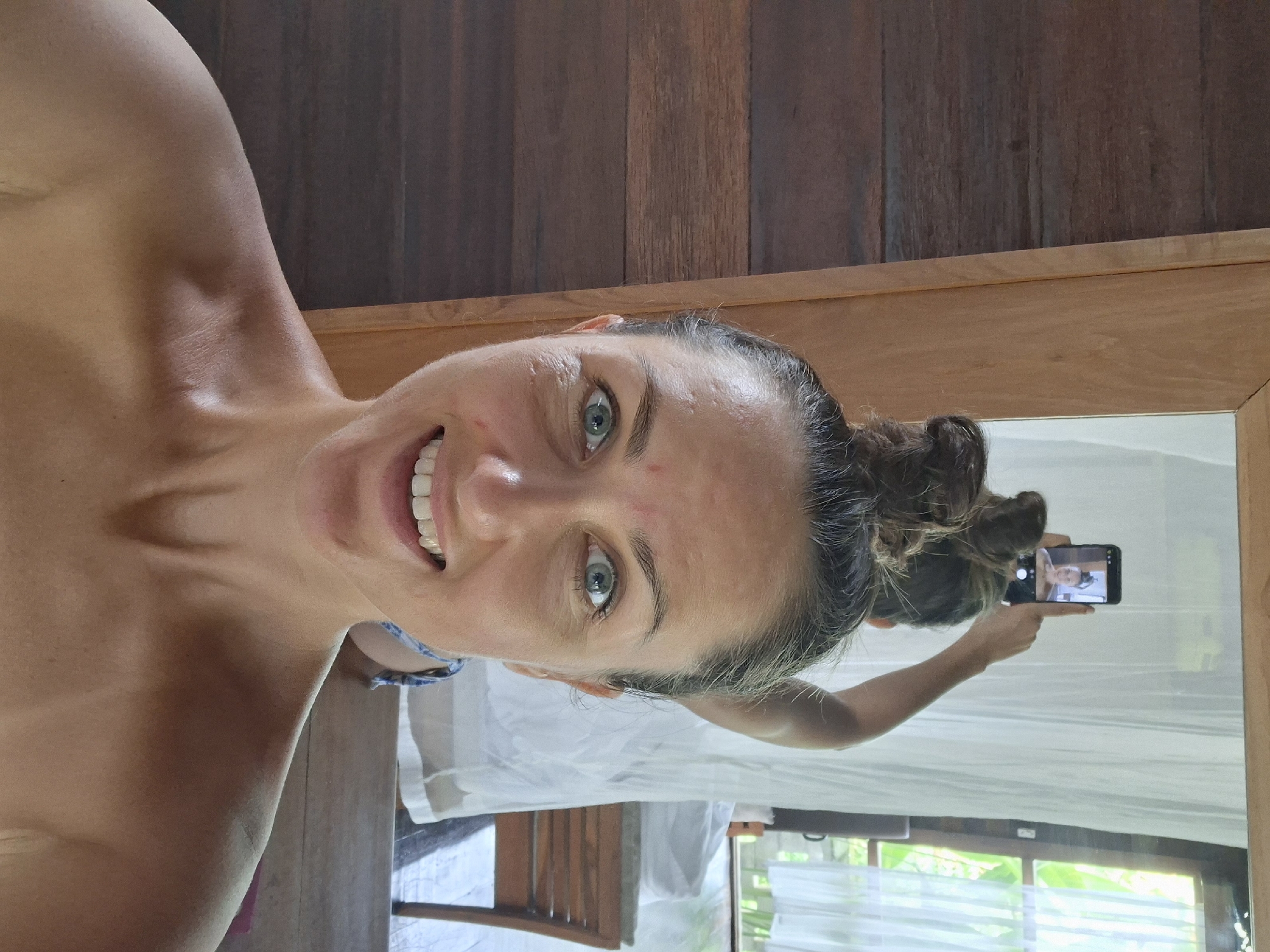 A woman with shoulder-length brown hair in a high bun smiles while taking a selfie in a mirror. She is fair-skinned with green eyes and visible freckles. She is topless and is likely in Ubud District, Indonesia, as suggested by the surrounding environment, which features a wooden wall, a wooden frame of the mirror, a white curtain, a wooden chair, and lush green foliage visible through a window. The reflection shows the woman holding a smartphone in her right hand to capture the selfie.