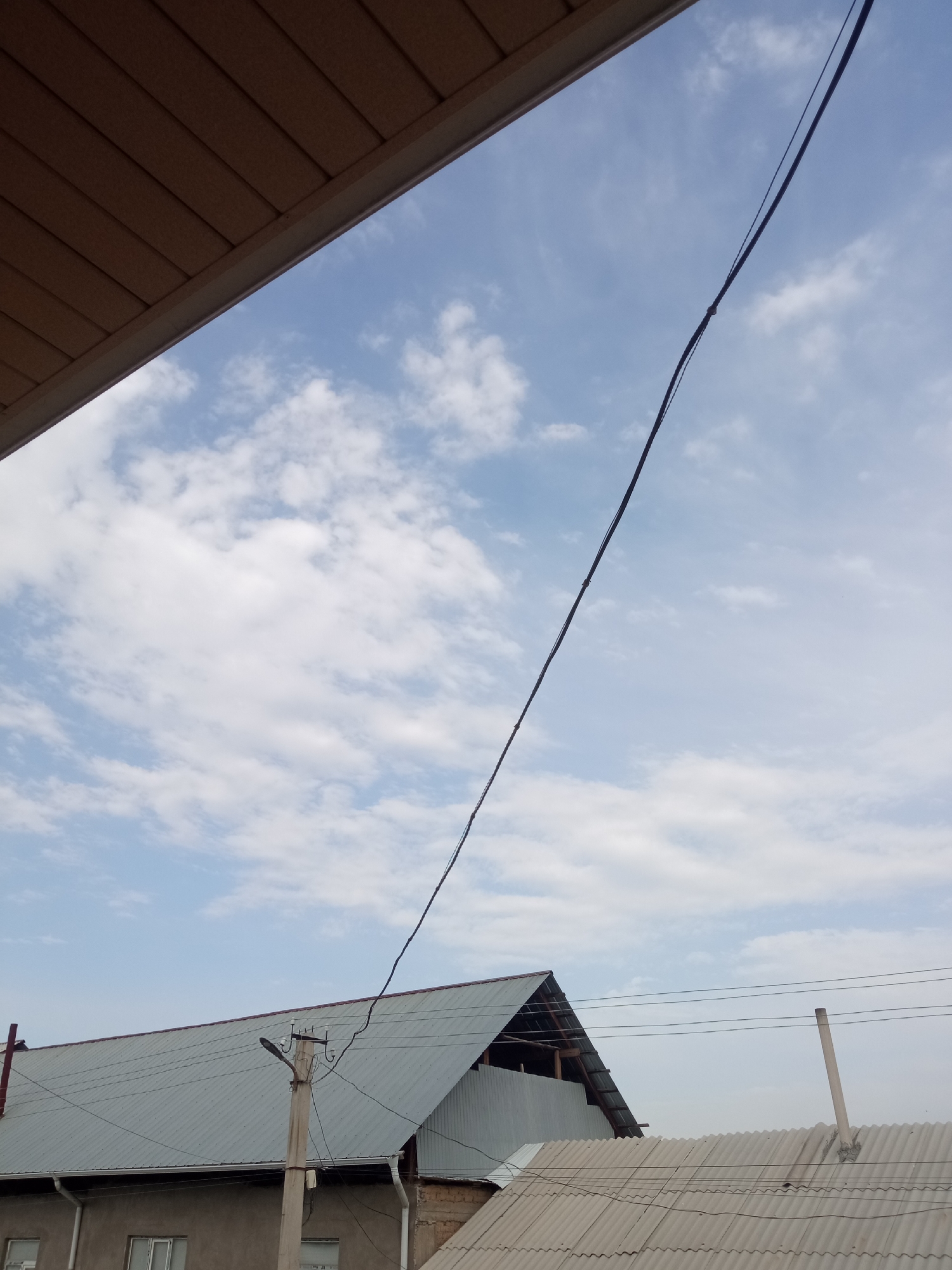 The image captures a view of the sky and rooftops in the city of Kirov, Kyrgyzstan. In the foreground, a brown soffit or eave extends from the top left corner, casting a shadow. A thin black wire, presumably an electrical cable, stretches diagonally across the frame from the upper right towards the lower left, supported by a concrete utility pole. The sky is a pale blue, dotted with scattered white clouds. Below the sky, the corrugated metal roofs of several buildings are visible. One prominent building has a gray, pitched roof with exposed wooden rafters underneath one section. To the right, another building has a lighter gray, corrugated roof. A concrete utility pole with insulators and wires stands near the center of the image, in front of the pitched roof. A partially visible building wall with windows is present in the lower left. The overall impression is a slightly overcast day in a residential or mixed-use area.