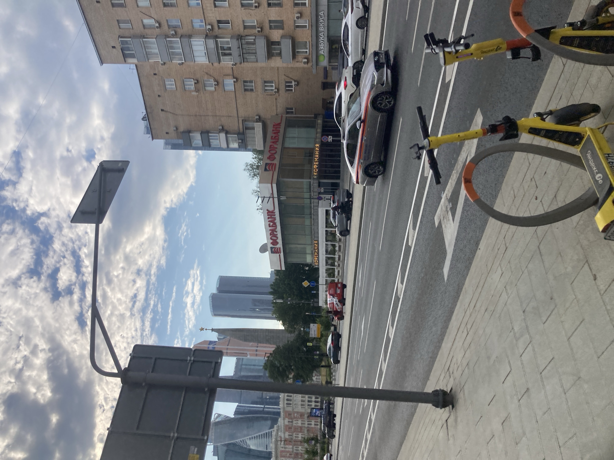 This is a full-frame, eye-level, outdoor, daytime photograph of a street scene in Moscow, Russia. In the foreground, two yellow electric scooters are parked on a paved sidewalk next to a street. The street is multi-lane with white lane markings. Cars are driving on the street, including a silver sedan with red stripes on its side, a white SUV, and a black sedan. In the mid-ground, a building with a glass facade and the word "ФОРБАНК" (FORABANK) written on it is visible, along with a sign that says "КОФЕМАНИЯ" (KOFEMANIA). To the right of the FORABANK building is a multi-story brick apartment building. Further in the background, several modern skyscrapers are visible, including the Moscow International Business Center skyscrapers. The sky is cloudy with patches of blue sky. A traffic sign is mounted on a pole in the left foreground.