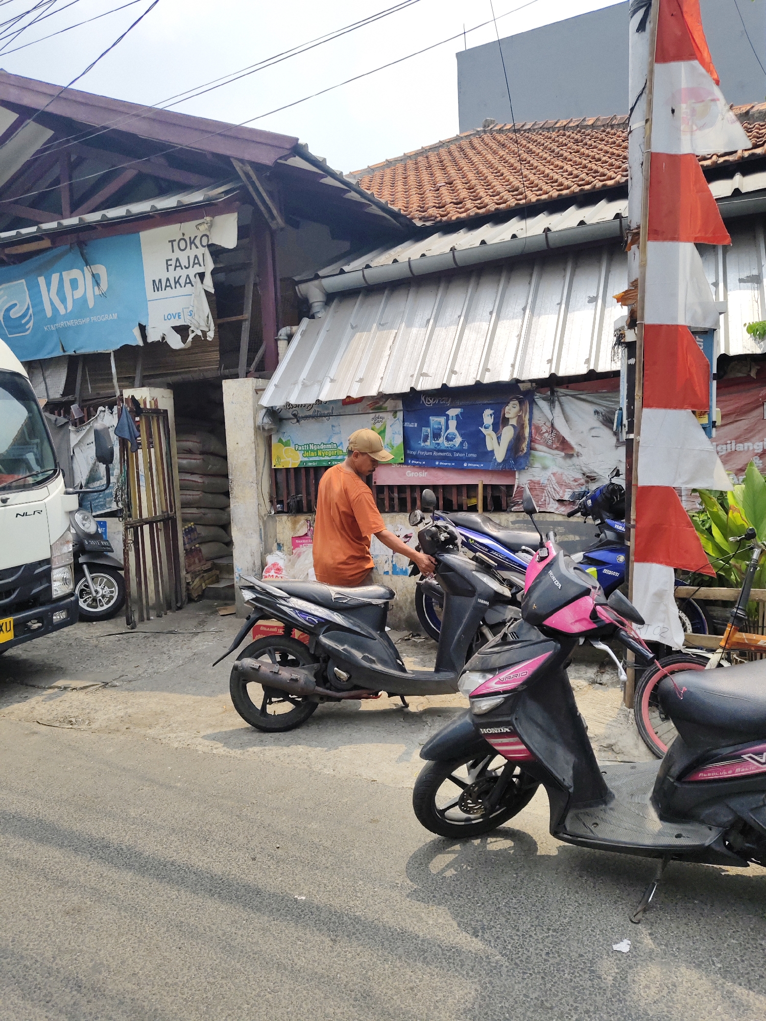 In South Jakarta, Indonesia, a man in an orange t-shirt and a tan cap stands next to a black motorcycle. He is holding onto the handlebars of the motorcycle and appears to be interacting with it. In the background, there are several other motorcycles parked in front of a building with a corrugated metal roof. A white truck is parked to the left of the scene. Several signs are visible on the building, including one that reads "KPP KT&PARTNERSHIP PROGRAM" and another with text in Indonesian that appears to be an advertisement. A red and white striped flag is also visible on a pole to the right. The ground is paved with asphalt and shows a shadow from overhead wires. The overall scene depicts a street-side area with parked vehicles and commercial establishments.