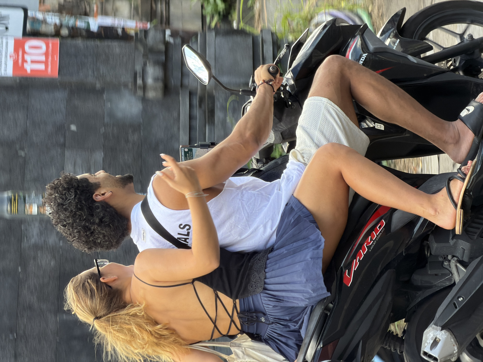 In North Kuta, Indonesia, a couple rides a black scooter. The woman, with blonde hair in a ponytail and wearing glasses, sits in front. She has on a black tank top with a lace-up back and a blue pleated skirt. A white tote bag hangs from her left shoulder. The man, with curly dark hair and a beard, drives the scooter. He is wearing a white tank top and white shorts. They are moving on a street with a gray textured wall behind them, and a red sign with "110" is visible on the wall. Green foliage lines the right side of the road, and a mirror is attached to the scooter.