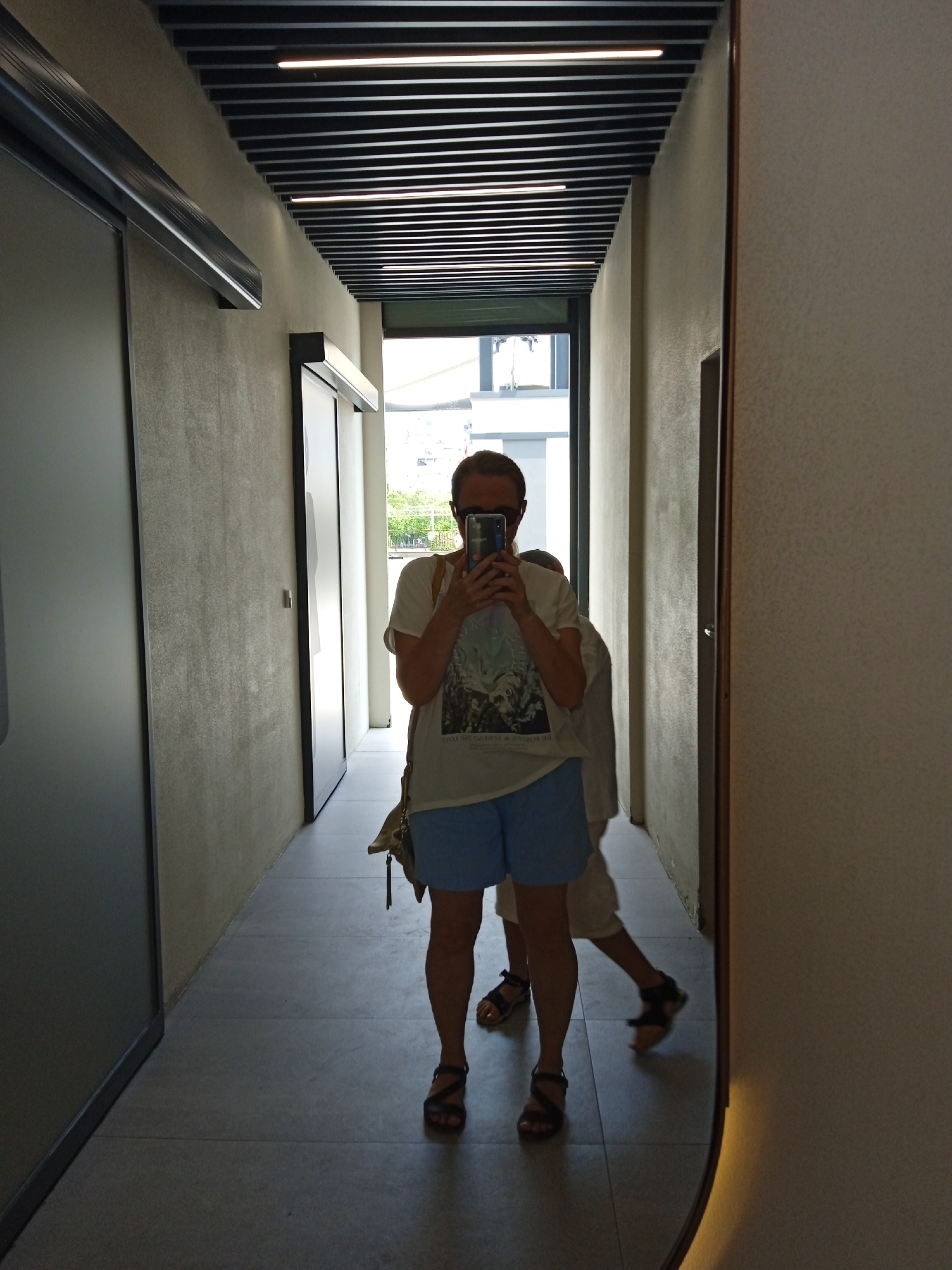 The image shows a woman taking a selfie in a hallway. She is wearing a white t-shirt, blue shorts, and sandals. She is holding a phone in her right hand and a bag is slung over her left shoulder. The hallway has a tiled floor and textured walls. A large mirror is on the right side of the hallway, reflecting the woman and another person in the background. The ceiling has a black slatted design with lights. The overall impression is one of a modern, indoor space. The location is identified as Mezitli, Turkey.