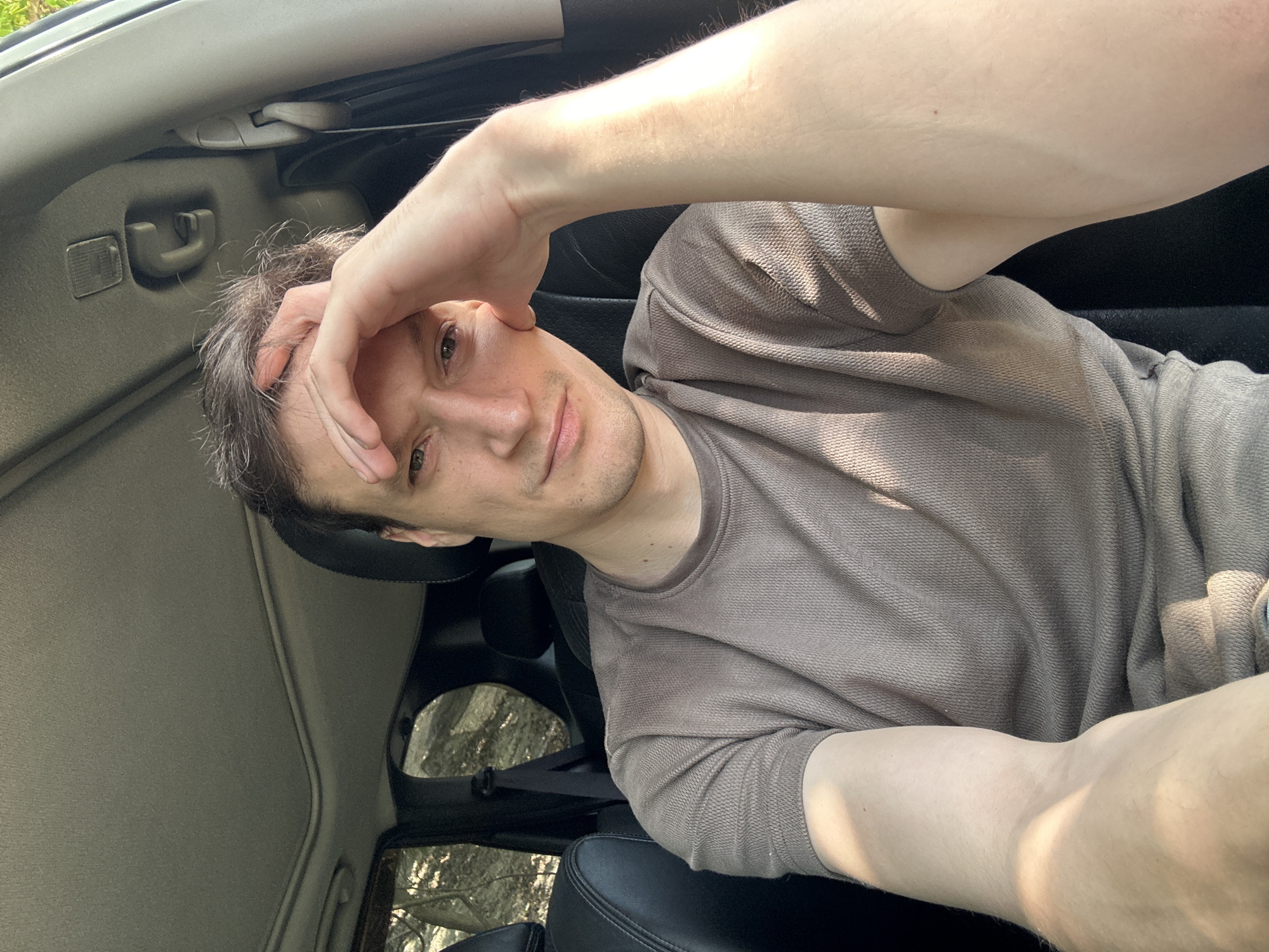 The image shows a close-up, selfie-style shot of a person sitting in the driver's seat of a car, likely in Makiv, Ukraine, given the context. The person has fair skin, brown hair, and is looking towards the camera with their right hand resting on their forehead. They are wearing a brown, textured t-shirt. The interior of the car is visible, including black leather seats, the car's headliner, and a portion of the sunroof. Outside the car windows, trees and possibly rocks are visible, suggesting a rural or wooded setting. The lighting indicates it is daytime, with sunlight casting shadows and illuminating parts of the person's arm and the car's interior.