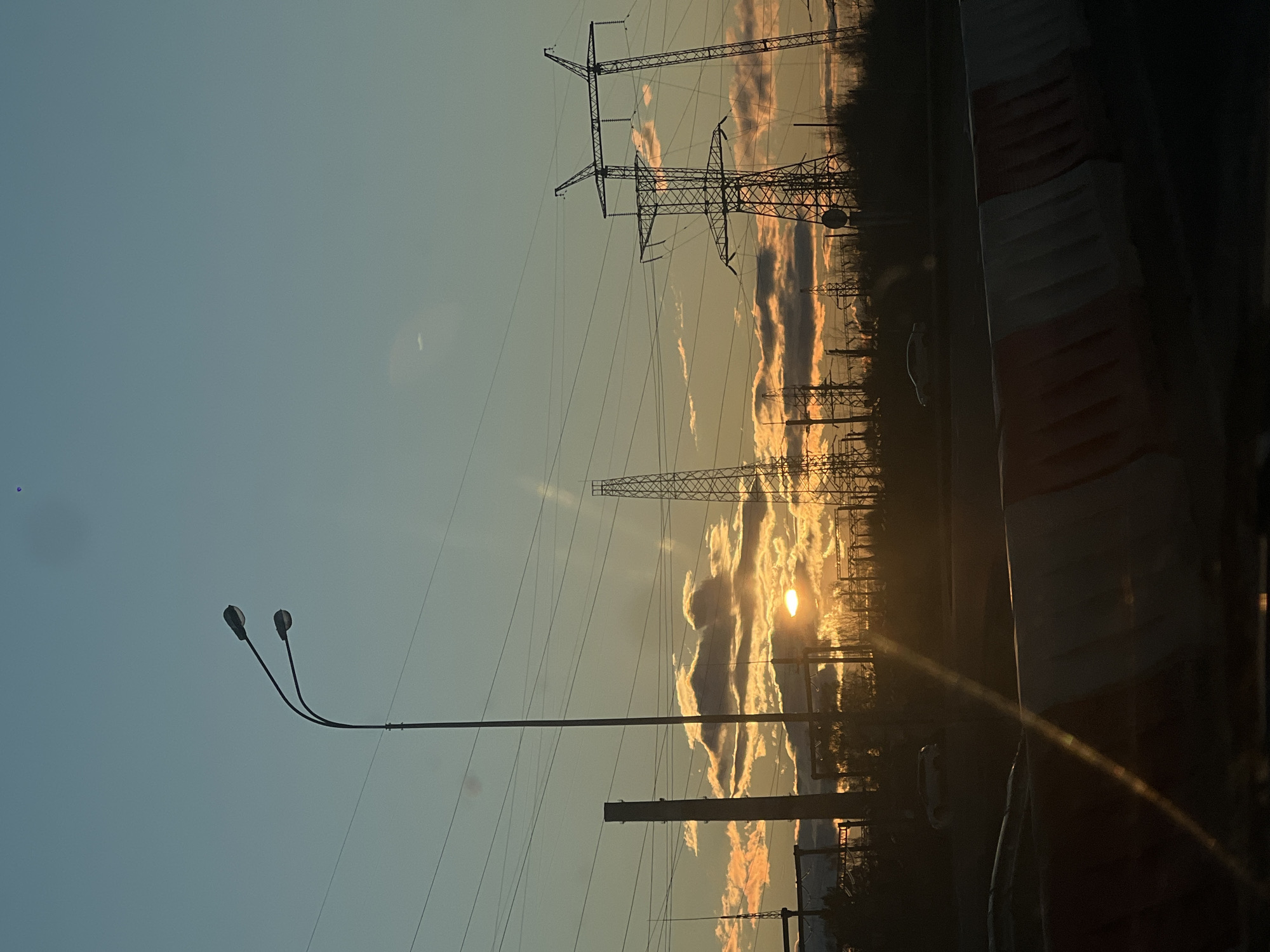 The image, correctly oriented horizontally, depicts an outdoor landscape scene during sunset or sunrise in Zelenodolsk, Russia. The sky occupies the upper two-thirds of the frame, transitioning from a pale blue at the top to warm orange and yellow hues near the horizon. The sun is prominently visible on the right side, low on the horizon, casting a bright glare and creating lens flares. Scattered clouds are illuminated from below, displaying warm orange and pink tones.
Several large electricity transmission pylons are silhouetted against the glowing sky, extending from the middle ground into the background. Numerous power lines stretch across the frame, connecting these pylons. In the left foreground, a modern street light with two curved arms and two lamps stands silhouetted against the bright sky. A dark, silhouetted line of trees or dense vegetation marks the distant horizon.
The lower part of the image is dark, suggesting ground level or a road. A faint white car can be discerned in the far distance on the right. On the extreme right foreground, a dark, vertical structure, possibly a barrier or part of a fence, is partially visible, showing hints of red and white stripes. No specific activity is taking place, and no visible text is present in the image.
